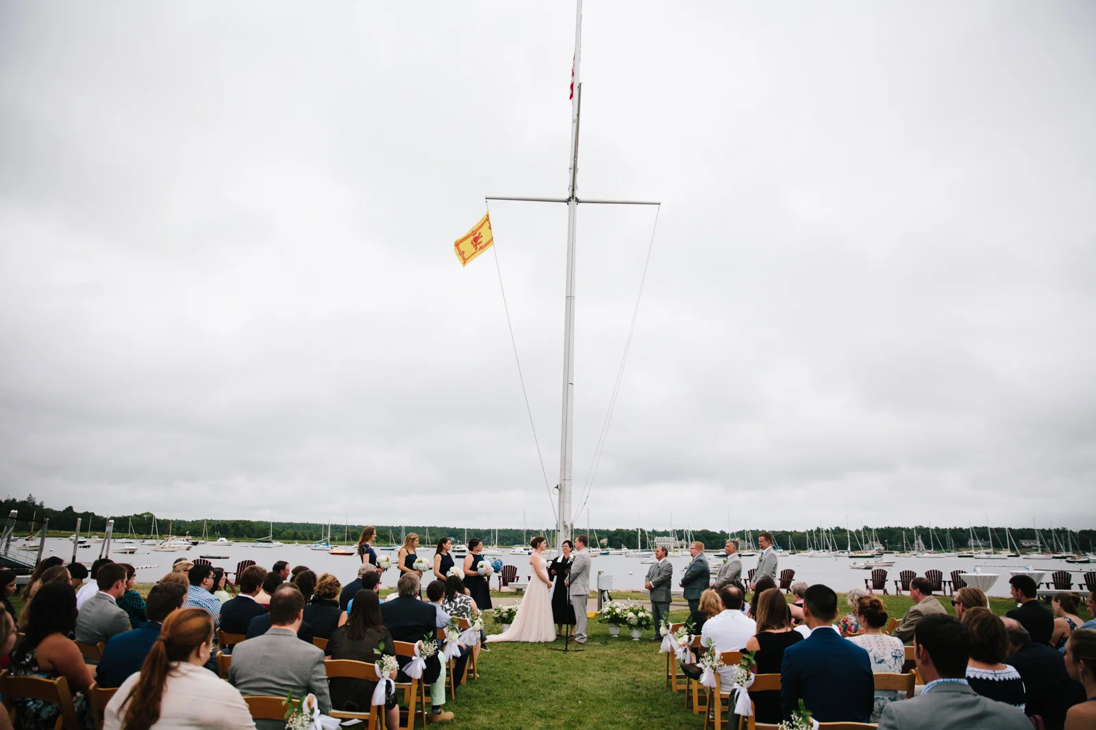 Outdoor wedding ceremony by the water with guests seated on wooden chairs, a bride and groom facing each other, and wedding party members standing nearby, under cloudy skies.