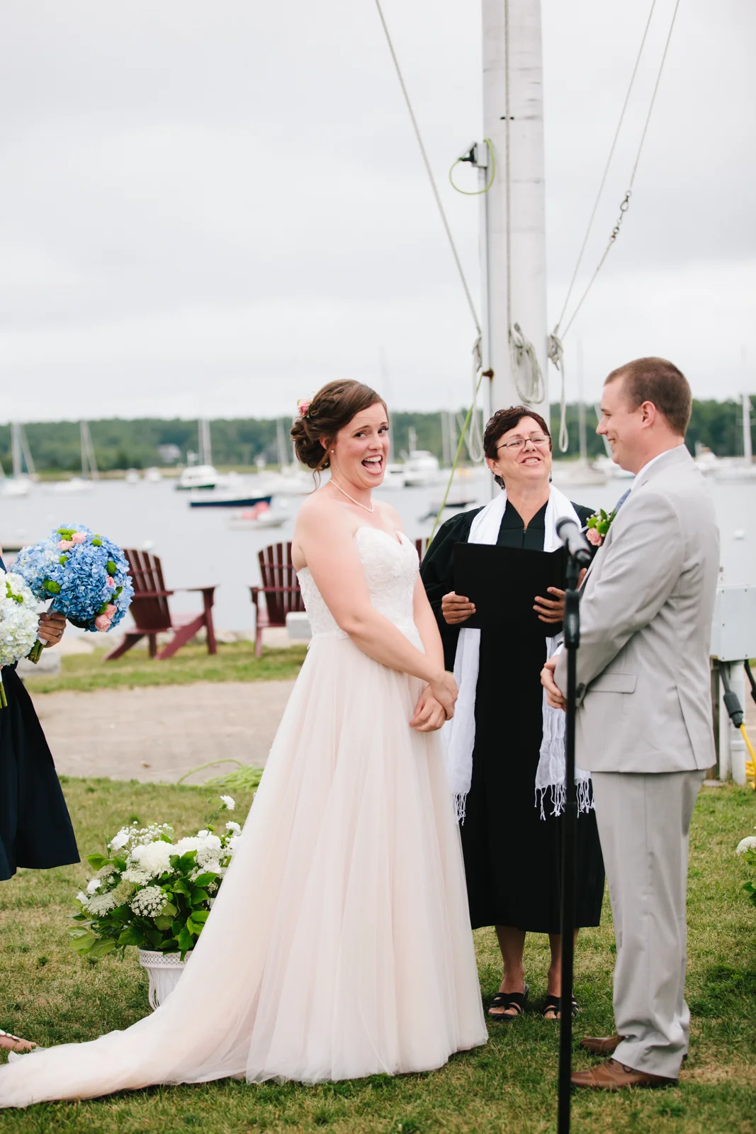 A wedding ceremony taking place outdoors near a marina with sailboats in the background. The bride, in a white strapless gown, is smiling and holding hands with the groom, who is dressed in a light gray suit. The officiant, standing between them, is 