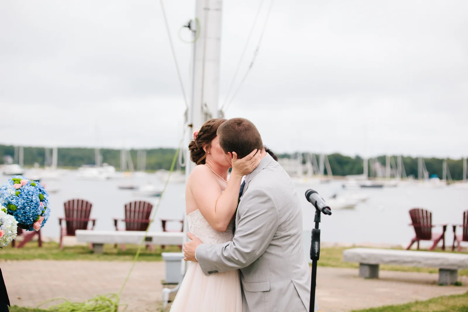 Bride and groom sharing a kiss outdoors near a waterfront with boats, during a wedding ceremony.