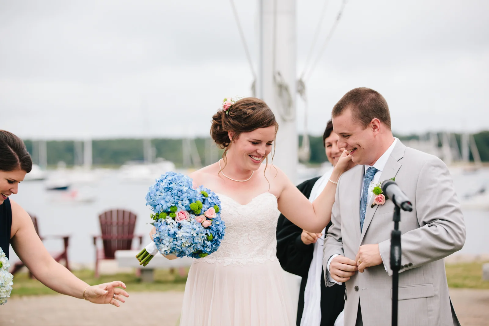 A bride and groom smiling at each other during their wedding ceremony near a marina with boats in the background. The bride holds a bouquet of blue and pink flowers, and the groom wears a light gray suit with a pink boutonniere.
