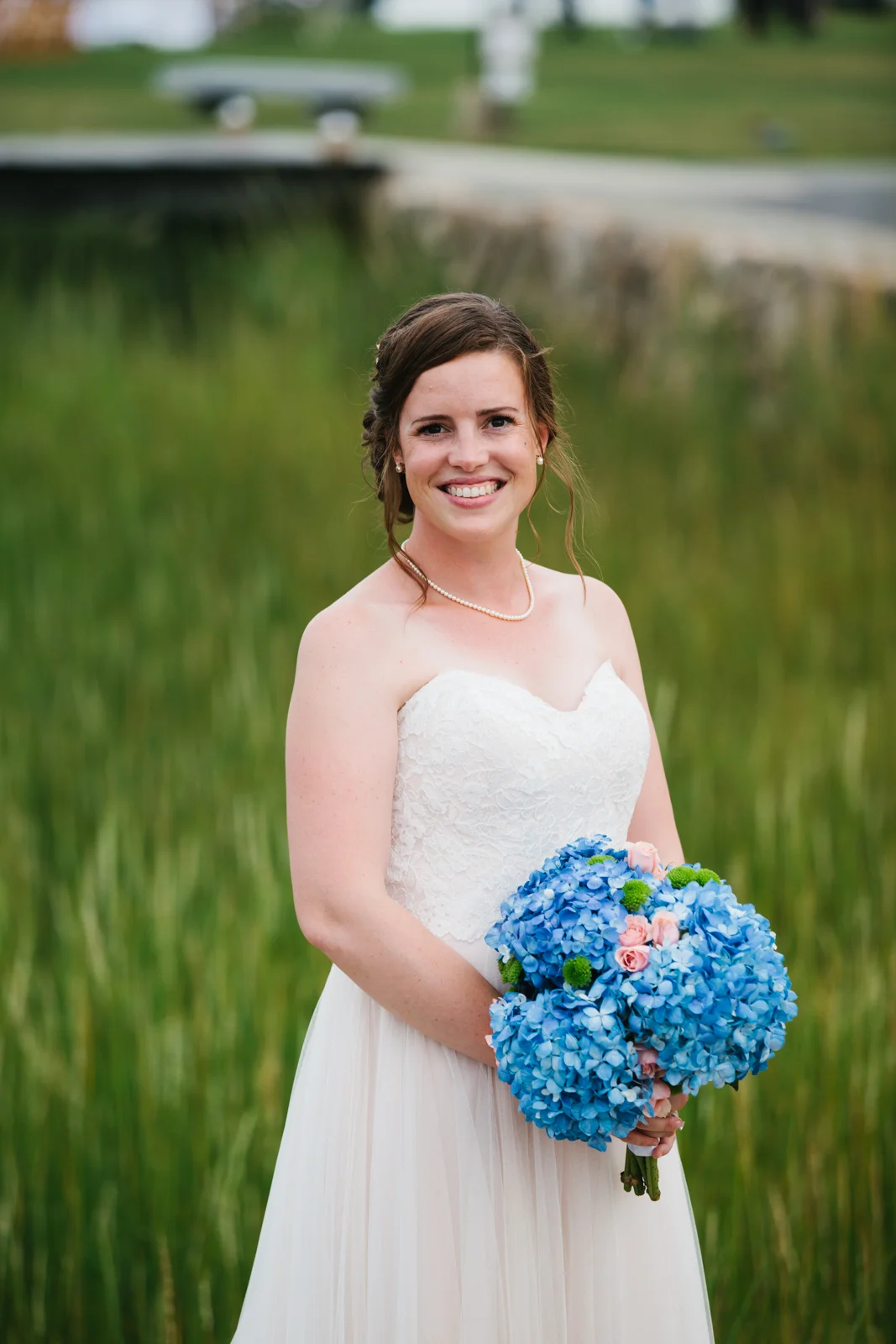 A smiling bride in a strapless white wedding dress holding a bouquet of blue hydrangeas and pink roses stands outdoors near a pond with green grass in the background.