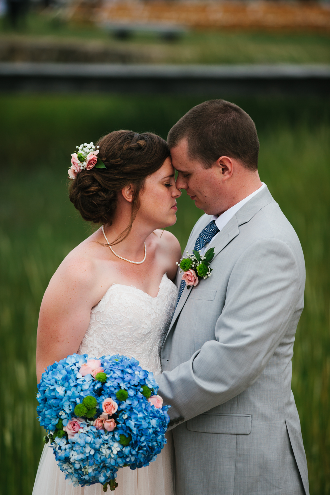 A bride and groom stand close with foreheads touching, holding a bouquet of blue hydrangeas with pink roses and green accents, outdoors near a green body of water.