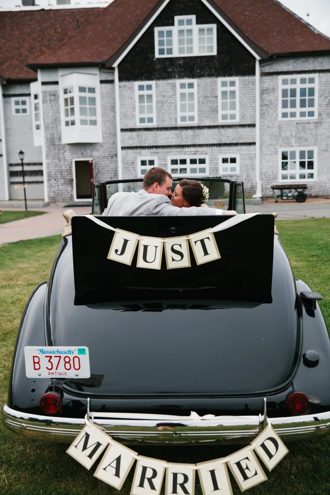 A newly married couple sitting in a vintage black convertible car, with wedding signs reading 'JUST MARRIED' attached to the back of the car, parked on a grassy area in front of a large house.