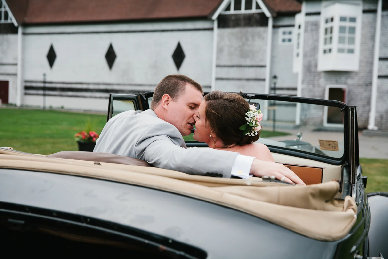 A newlywed couple in wedding attire sharing an intimate moment in a vintage convertible car outside a house.