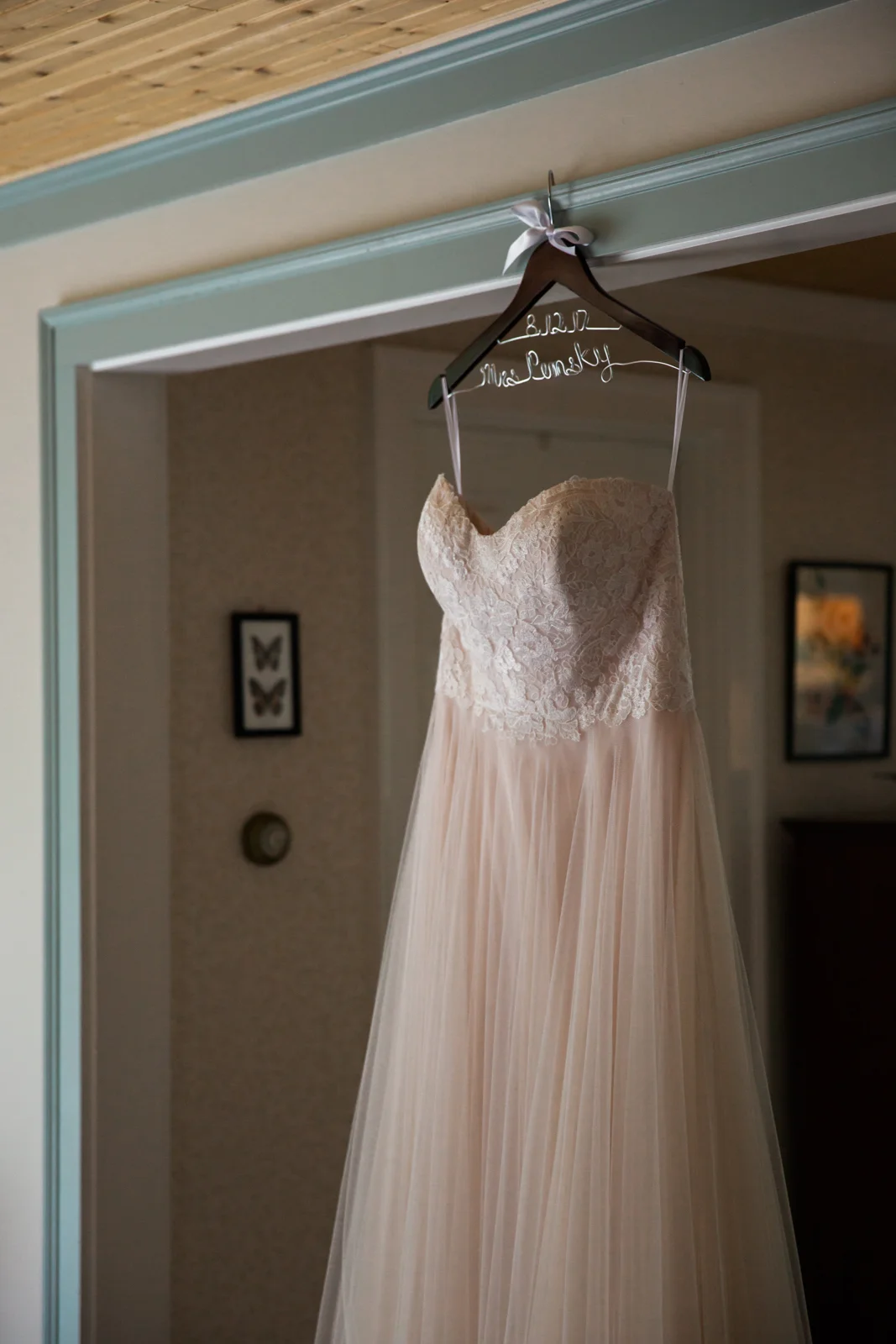 A wedding dress with a lace bodice and flowing tulle skirt hangs on a custom hanger with the words "Mrs. McFadden" and the date "8-12" written on it, inside a doorway.