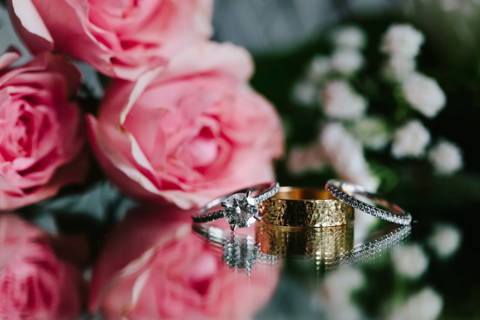 Close-up of a diamond engagement ring with a pink rose and a wedding band with small diamonds, reflected on a mirror surface, with blurred pink roses and white flowers in the background.
