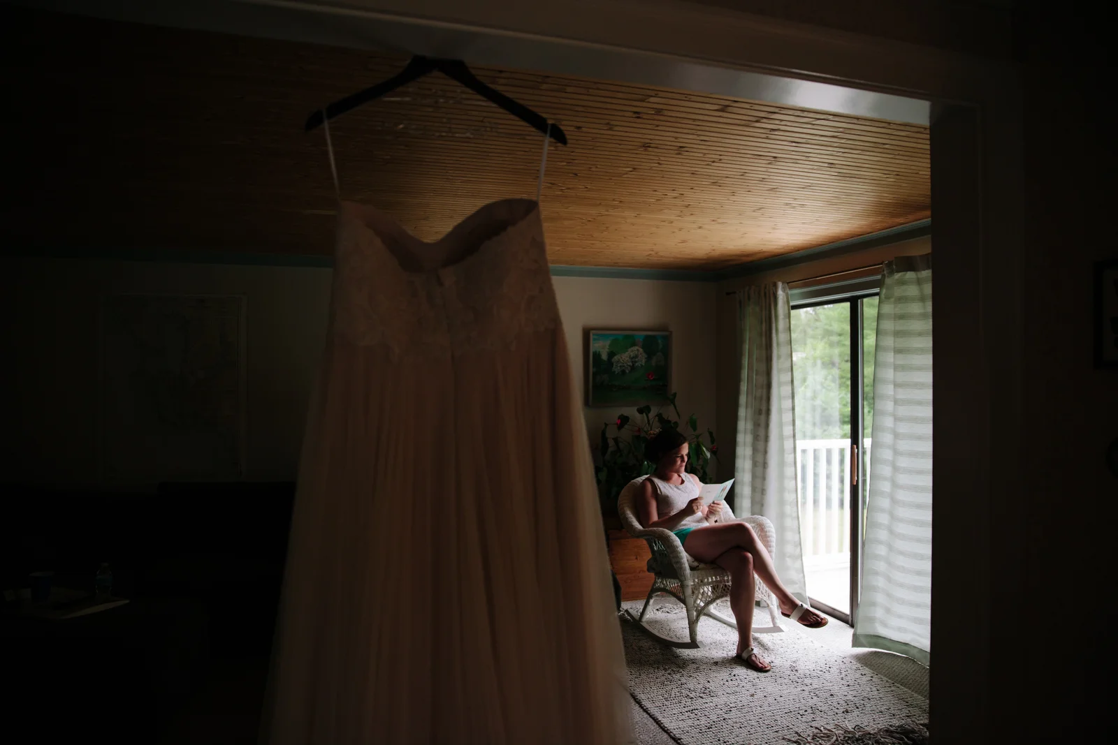 A woman sitting in a wicker chair reading a paper or a book near a glass door with white curtains, with a white dress hanging on a hanger in the foreground.