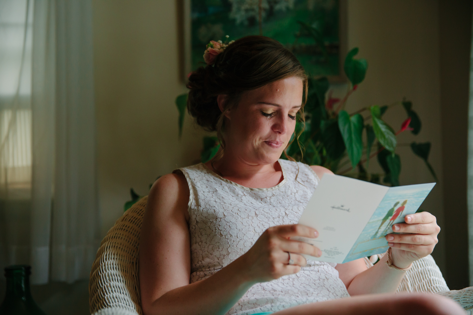 A woman with brown hair styled in an updo, wearing a sleeveless lace top, sitting in a wicker chair, reading a letter or card in a cozy, well-lit room with houseplants and artwork in the background.