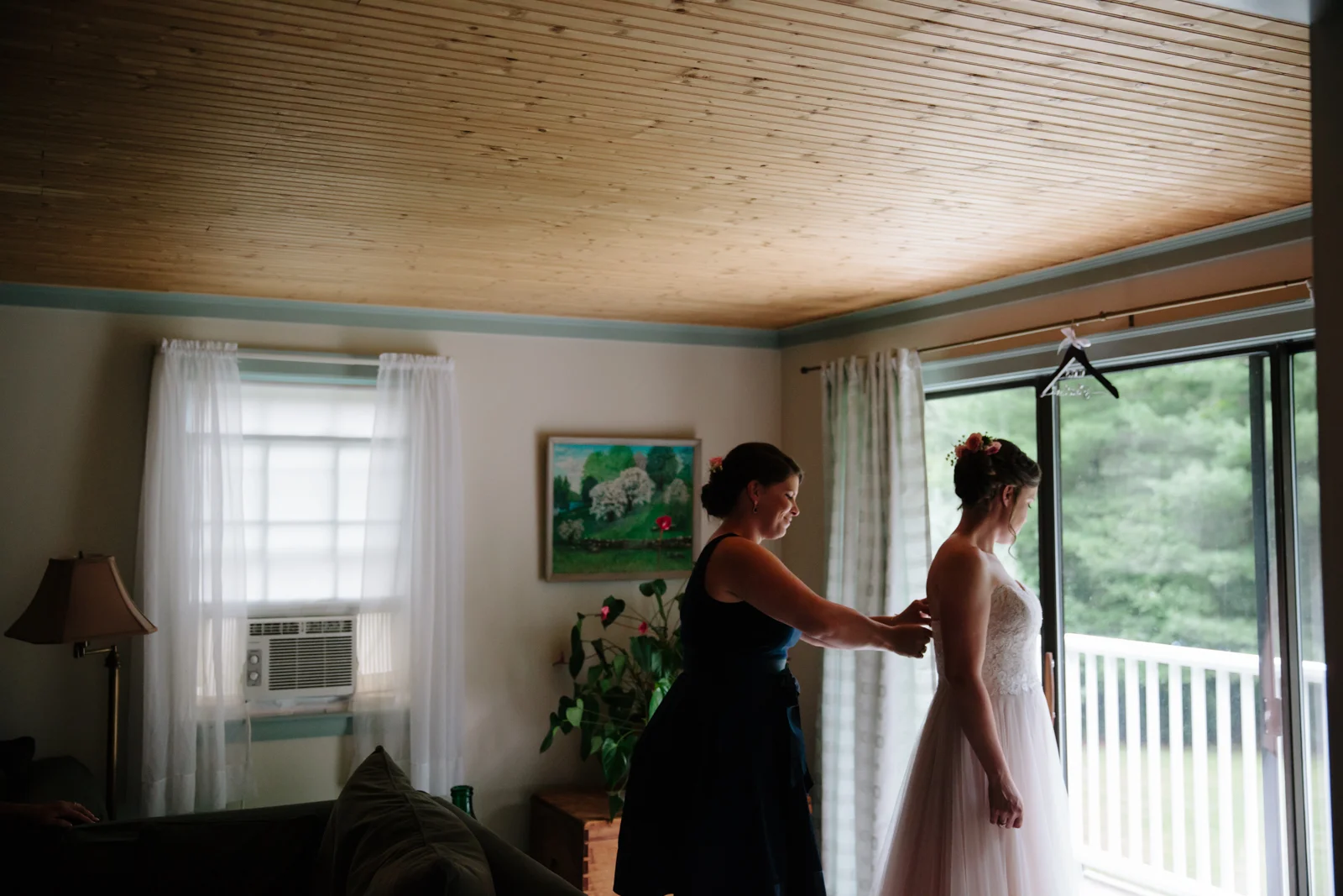 A woman in a wedding dress being assisted by another woman in a living room with windows and curtains, by a door leading to a balcony.