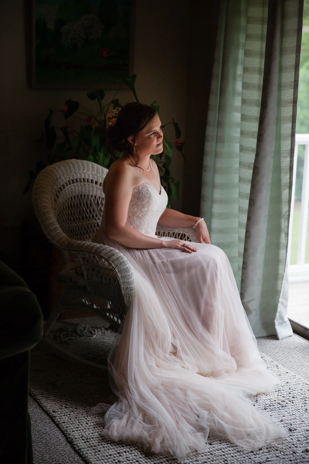 A bride sitting on a wicker chair indoors near a window, wearing a strapless wedding gown with a laced bodice and flowing skirt, looking contemplative.