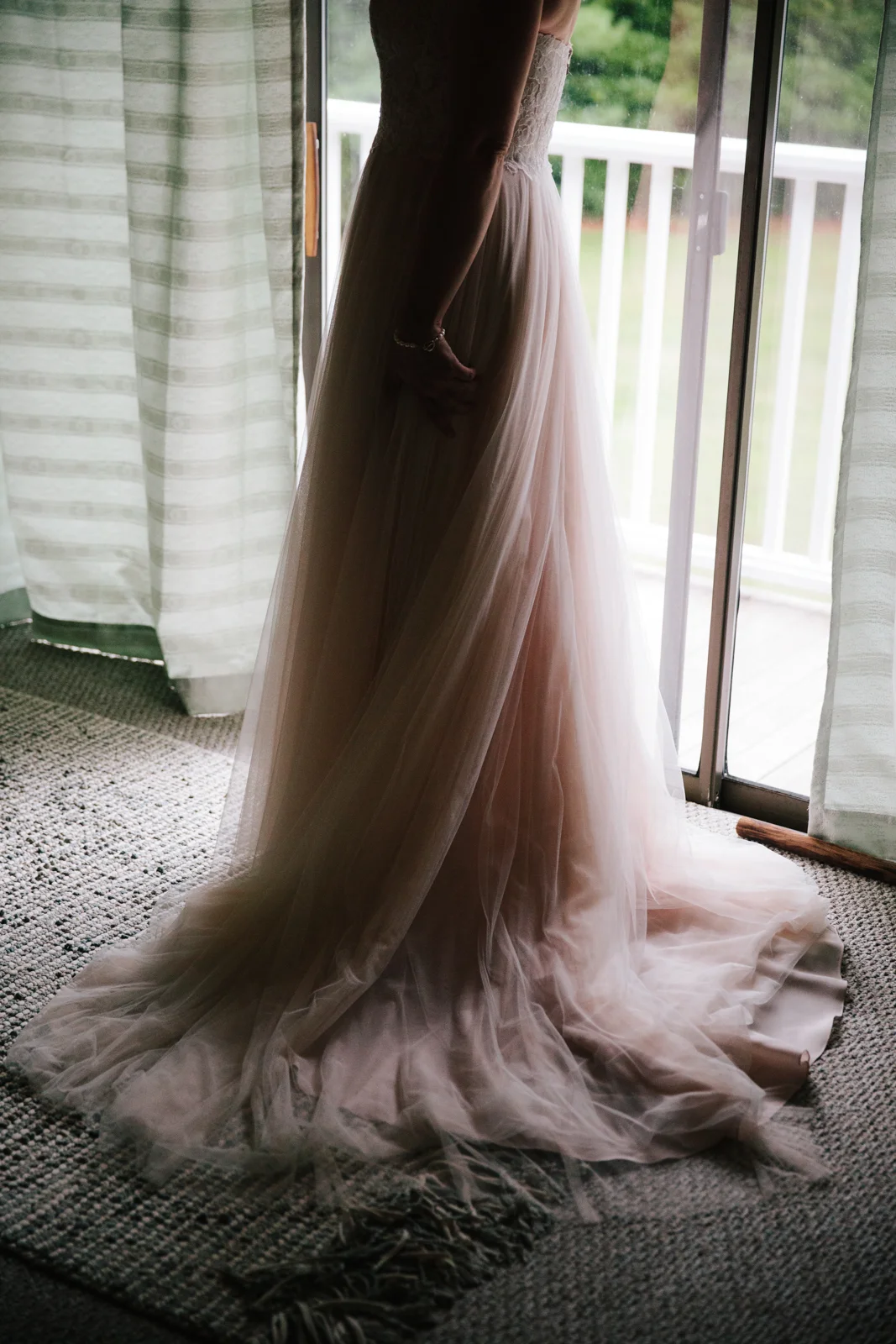 A woman in a wedding dress standing near a sliding glass door, with sunlight illuminating the dress and a view of greenery outside.