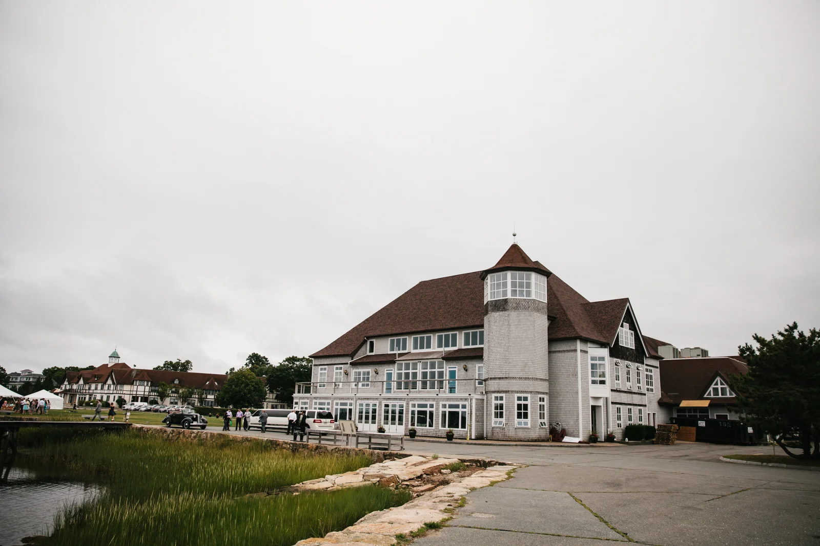 A large, historic-style building with a distinctive round tower and steep roof, situated near a pond, with people and cars visible in the nearby parking area.