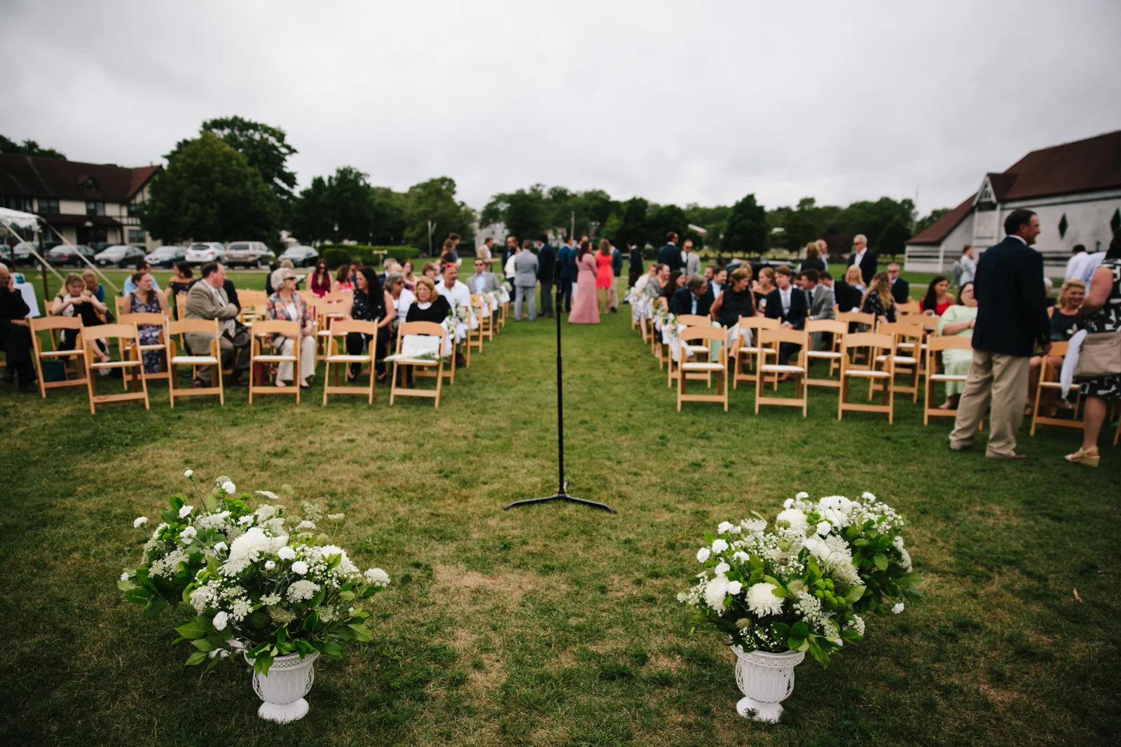 Outdoor wedding ceremony setup with wooden chairs arranged in rows on grass, decorated with white floral arrangements in urns at the front, under a cloudy sky, with guests seated and standing, and a microphone stand in the center.