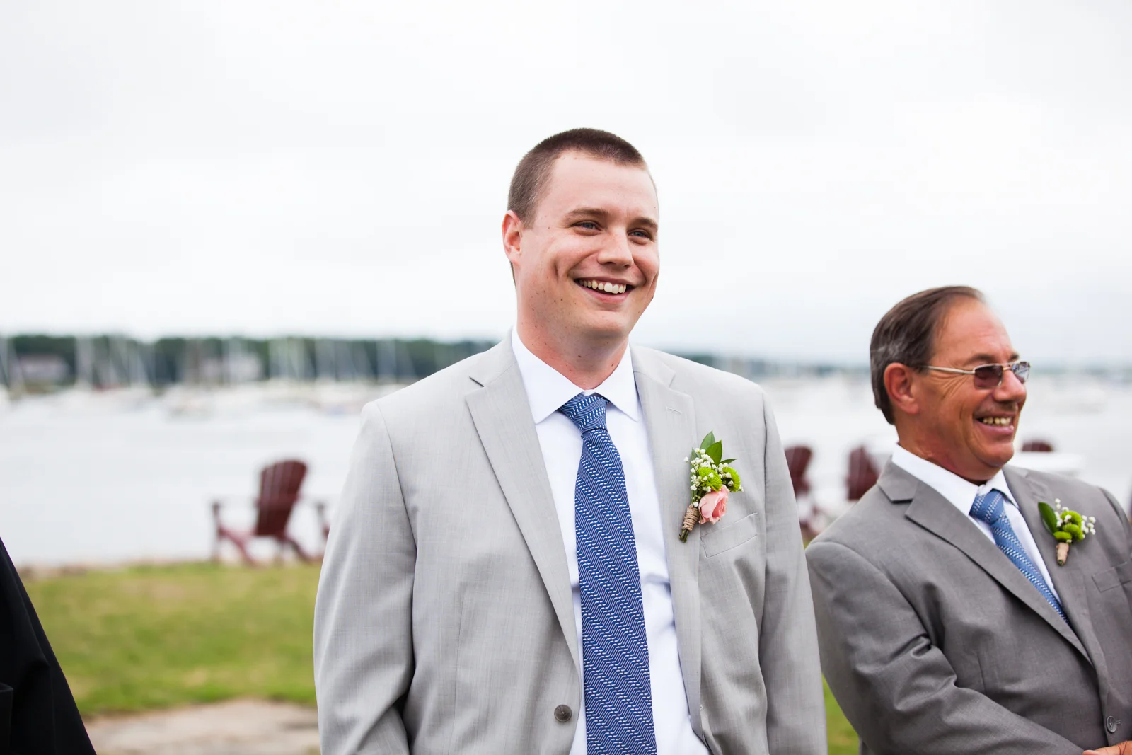 Two men dressed in suits at an outdoor wedding by a waterfront, smiling, with chairs and sailboats in the background.