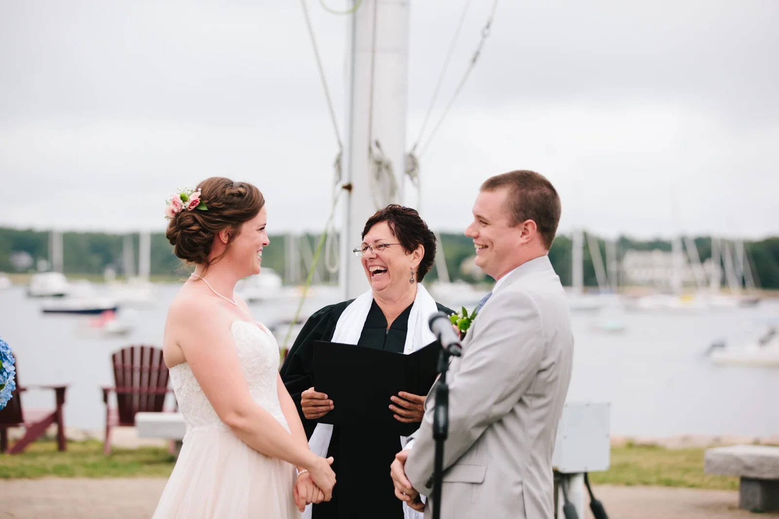 A bride and groom smiling at each other during their wedding ceremony outdoors with a woman officiant laughing between them, near a harbor with boats and overcast sky in the background.