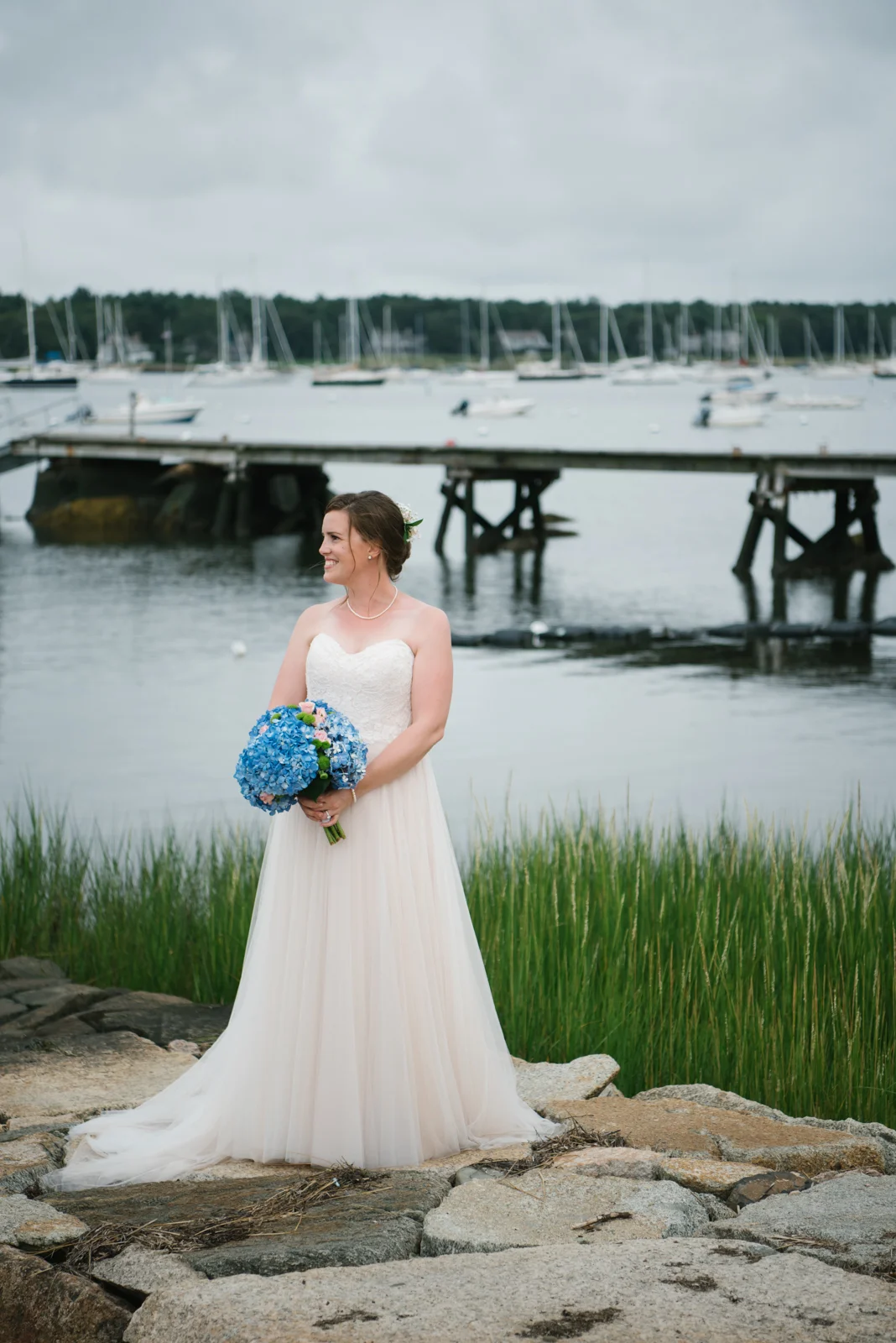 A bride in a white wedding dress holding a bouquet of blue and pink flowers, standing on rocks near a body of water with a dock and sailboats in the background.
