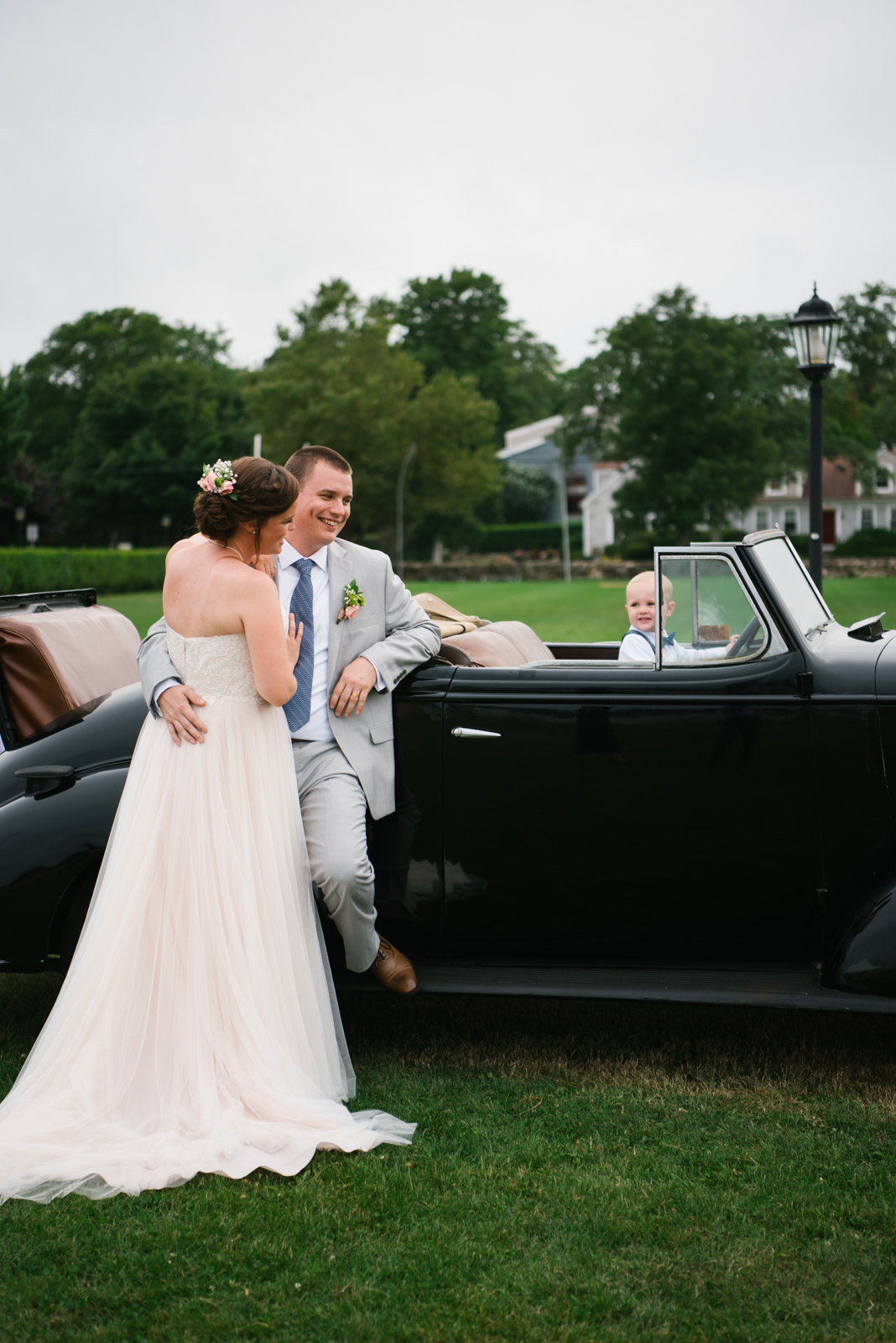 Bride and groom smiling and embracing next to a vintage black convertible car, with a young boy sitting inside the car, on a grassy lawn with trees and houses in the background.