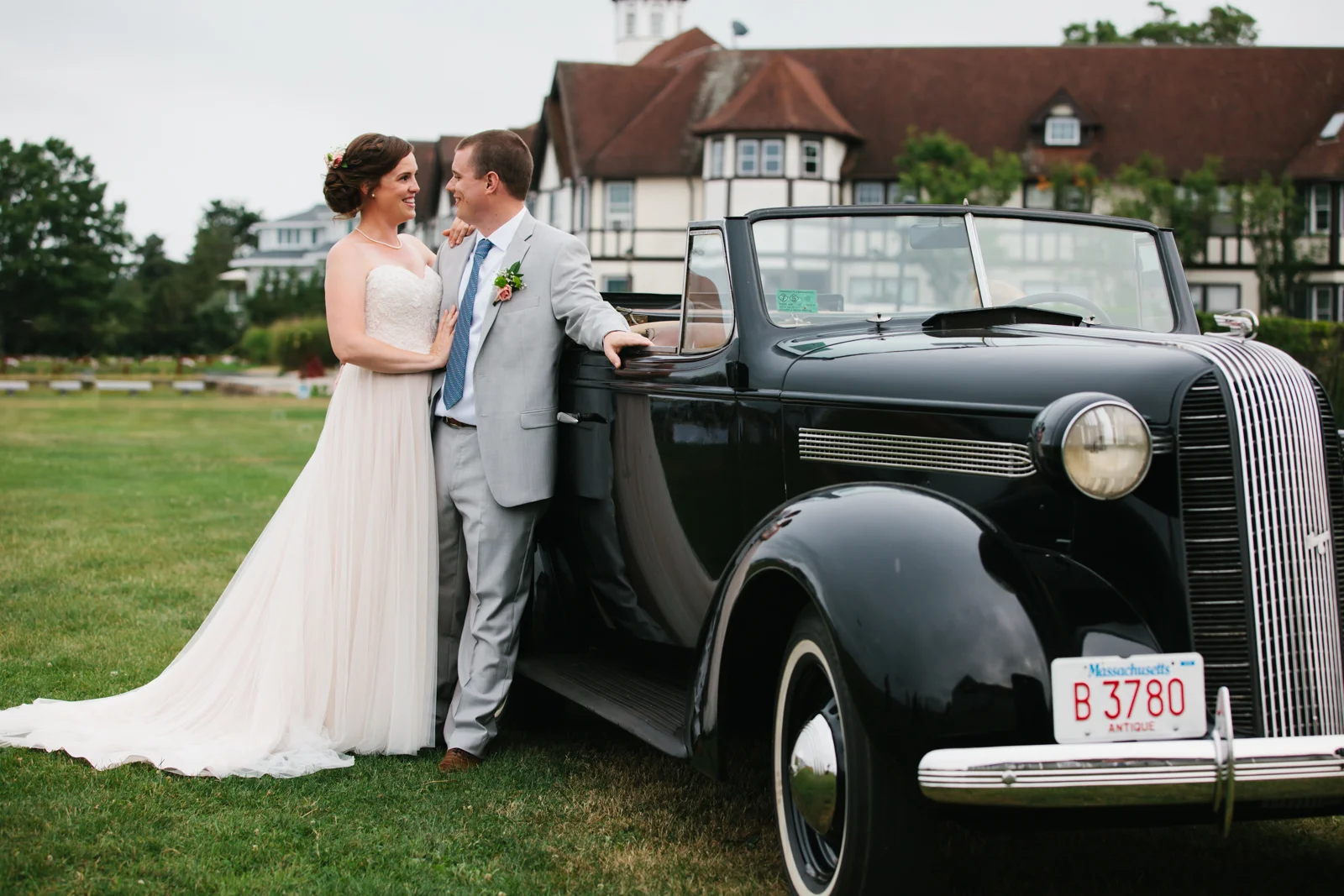 A bride and groom in wedding attire standing next to a vintage black car on a lawn with a large house in the background.