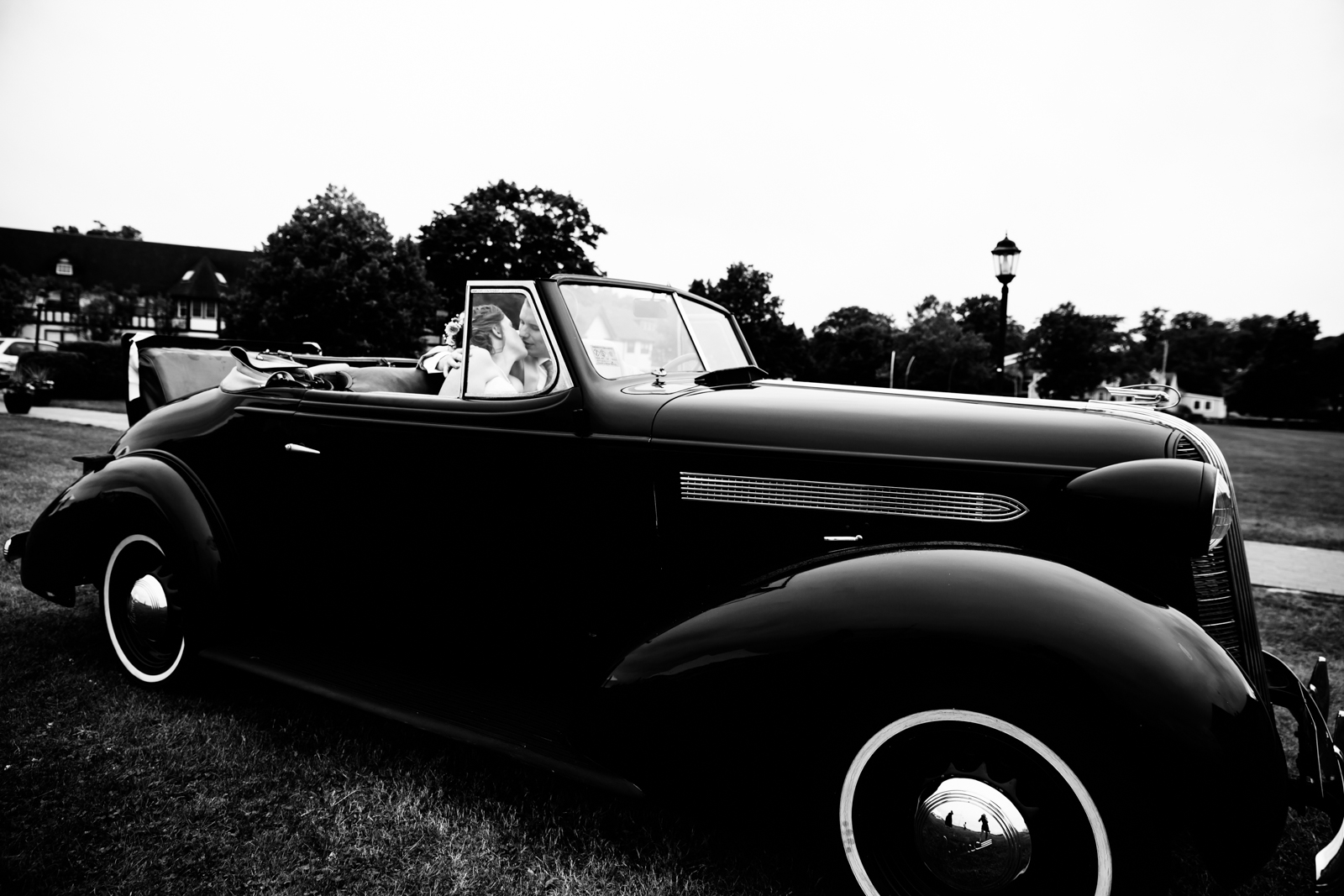 A vintage black convertible car parked on grass with a bride and groom inside, sharing a kiss. The scene appears to be at a wedding, with trees and a streetlamp in the background, captured in black and white.