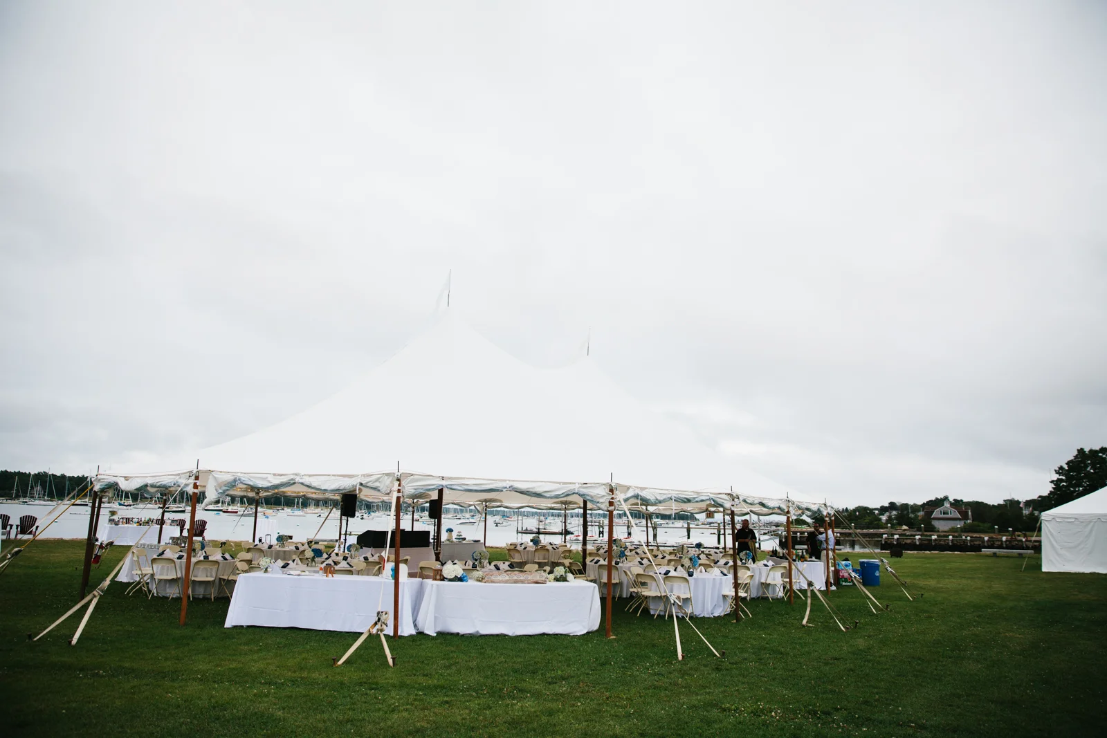 Outdoor event setup with white tents and tables on a grassy area near a marina with boats, under an overcast sky.
