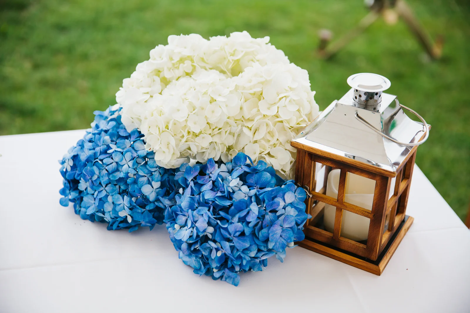 White and blue hydrangea flowers with a wooden candle lantern on a white table outdoors.