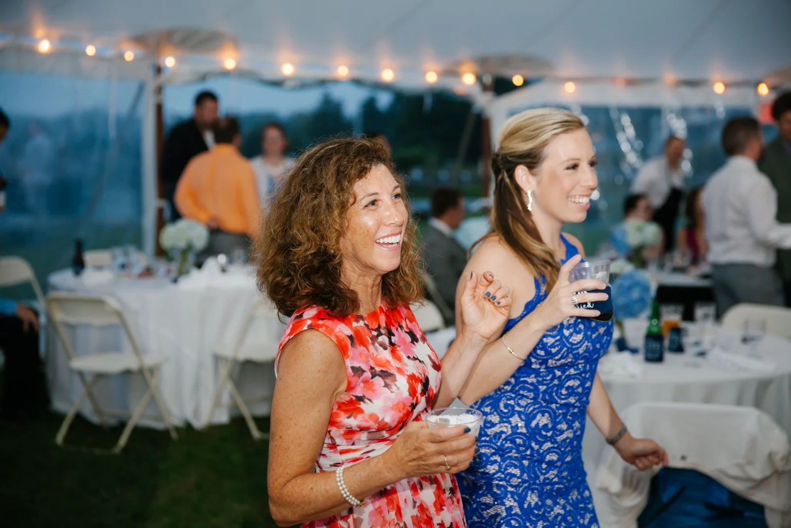 Two women enjoying a social event, smiling and holding drinks under a tent with string lights, surrounded by seated guests and tables.