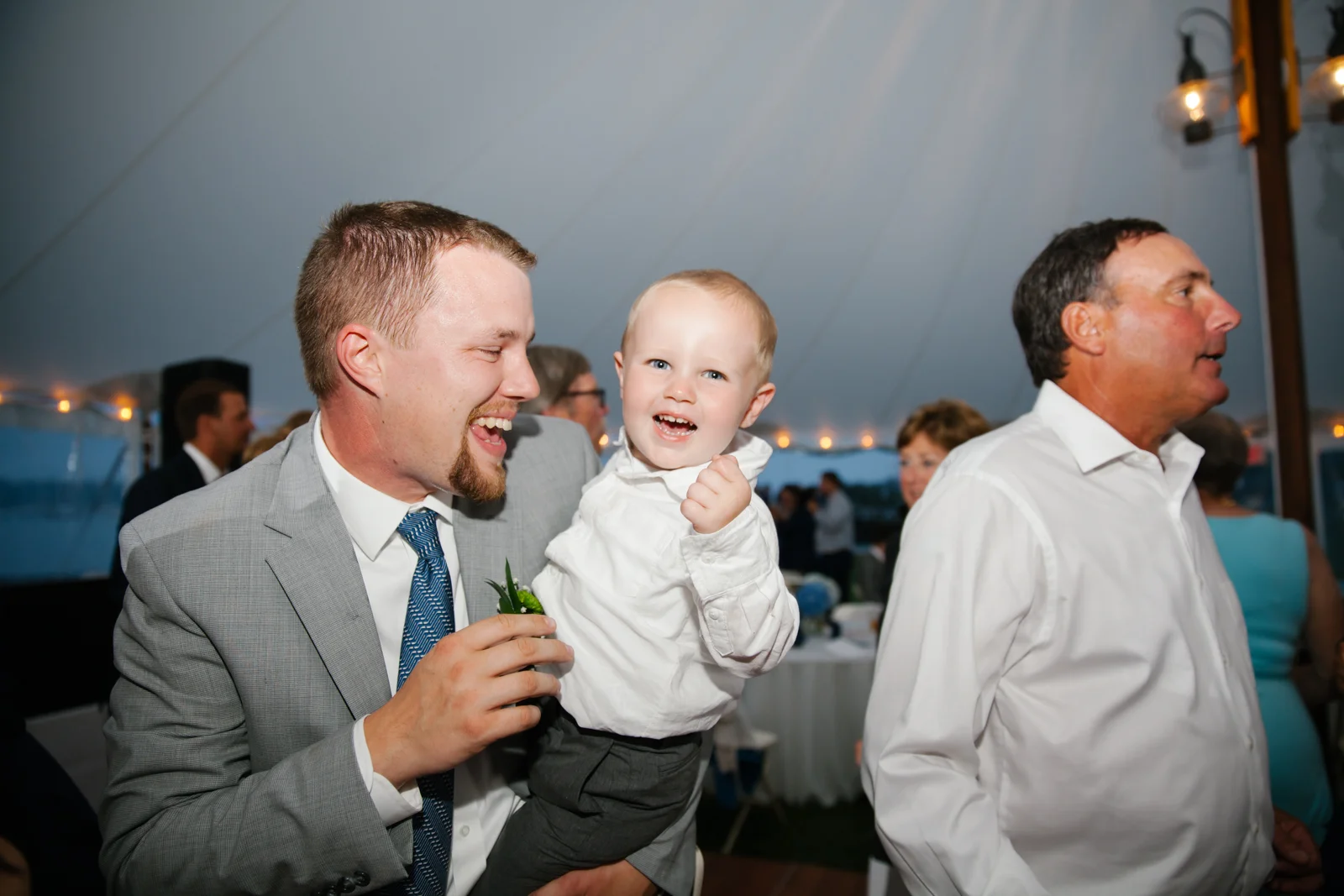 A man in a gray suit holding a smiling young boy in a white shirt at a wedding reception under a large tent, with other guests in the background.