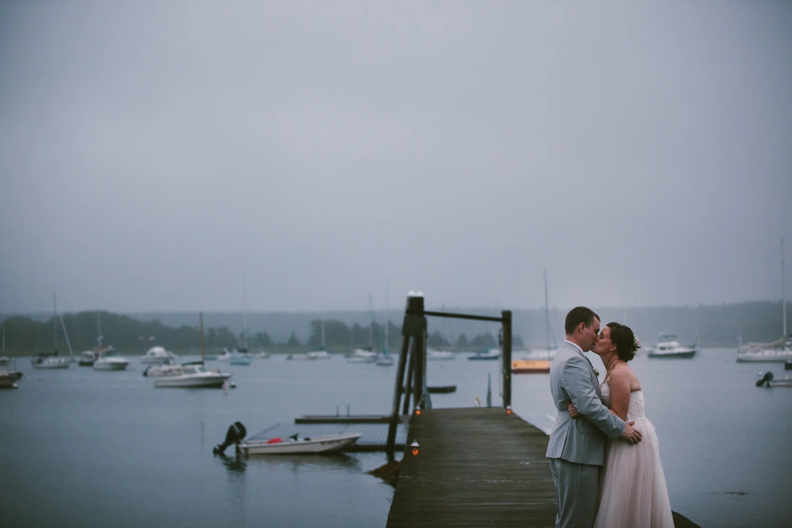 A newlywed couple sharing a kiss on a dock near boats on a cloudy day.