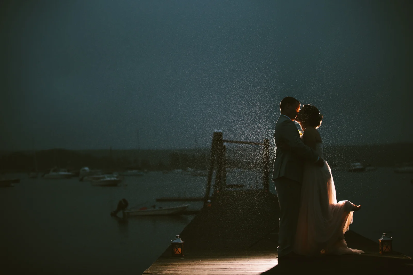 A couple in wedding attire embracing on a dock during sunset at a marina, with boats in the background and gentle lighting