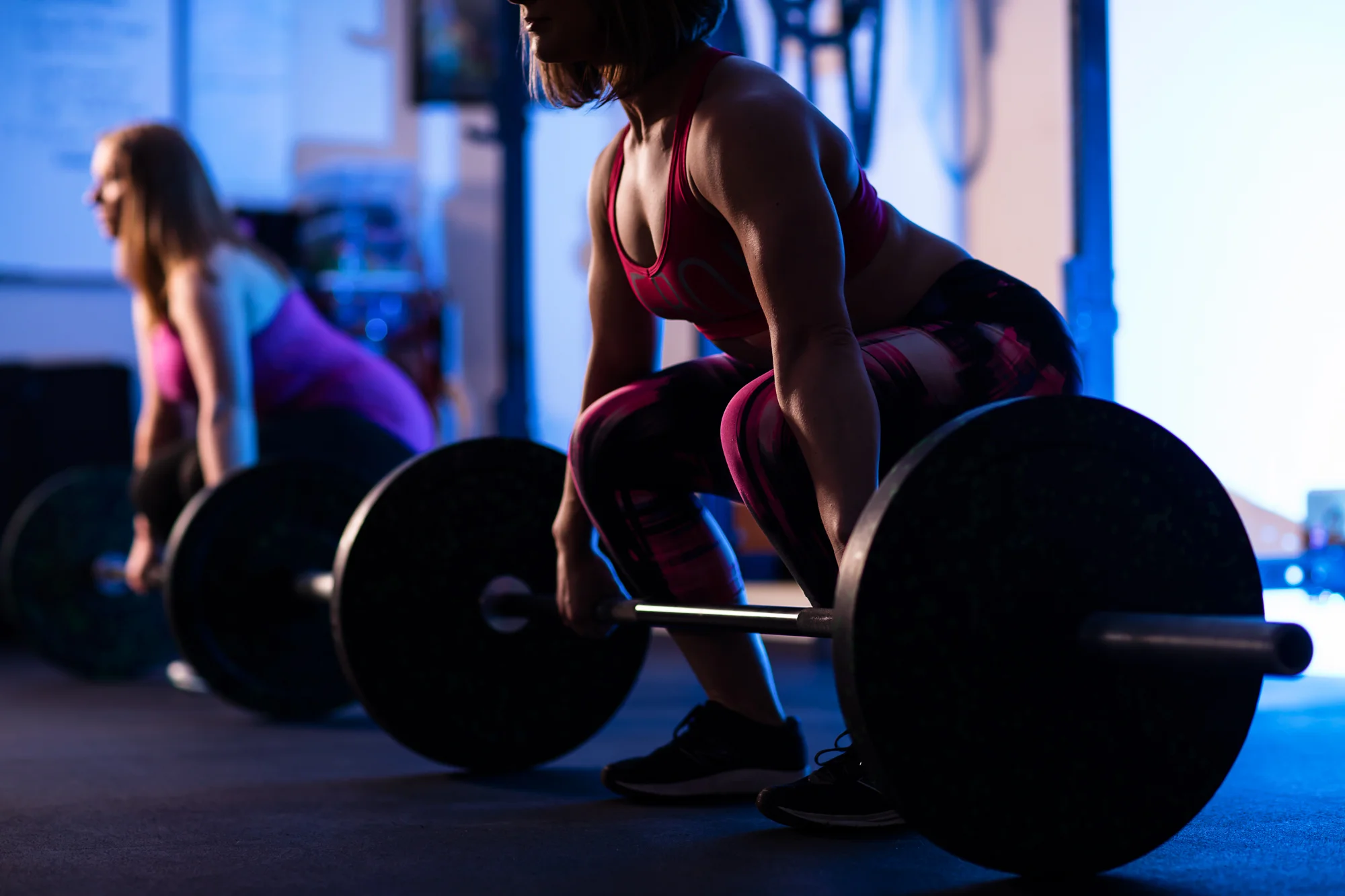 Two women in workout attire lifting barbells in a gym with dim lighting and blue hue.