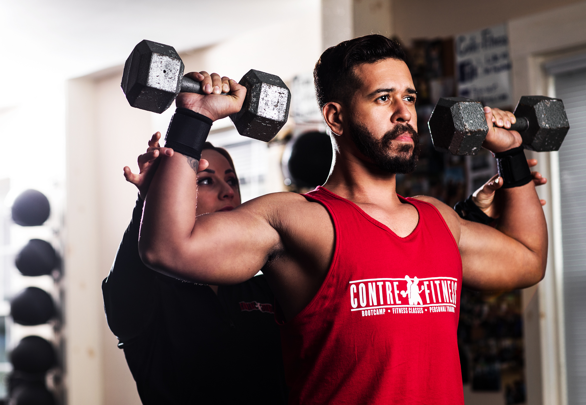Man doing a shoulder exercise with dumbbells in a gym, with a trainer assisting in the background.