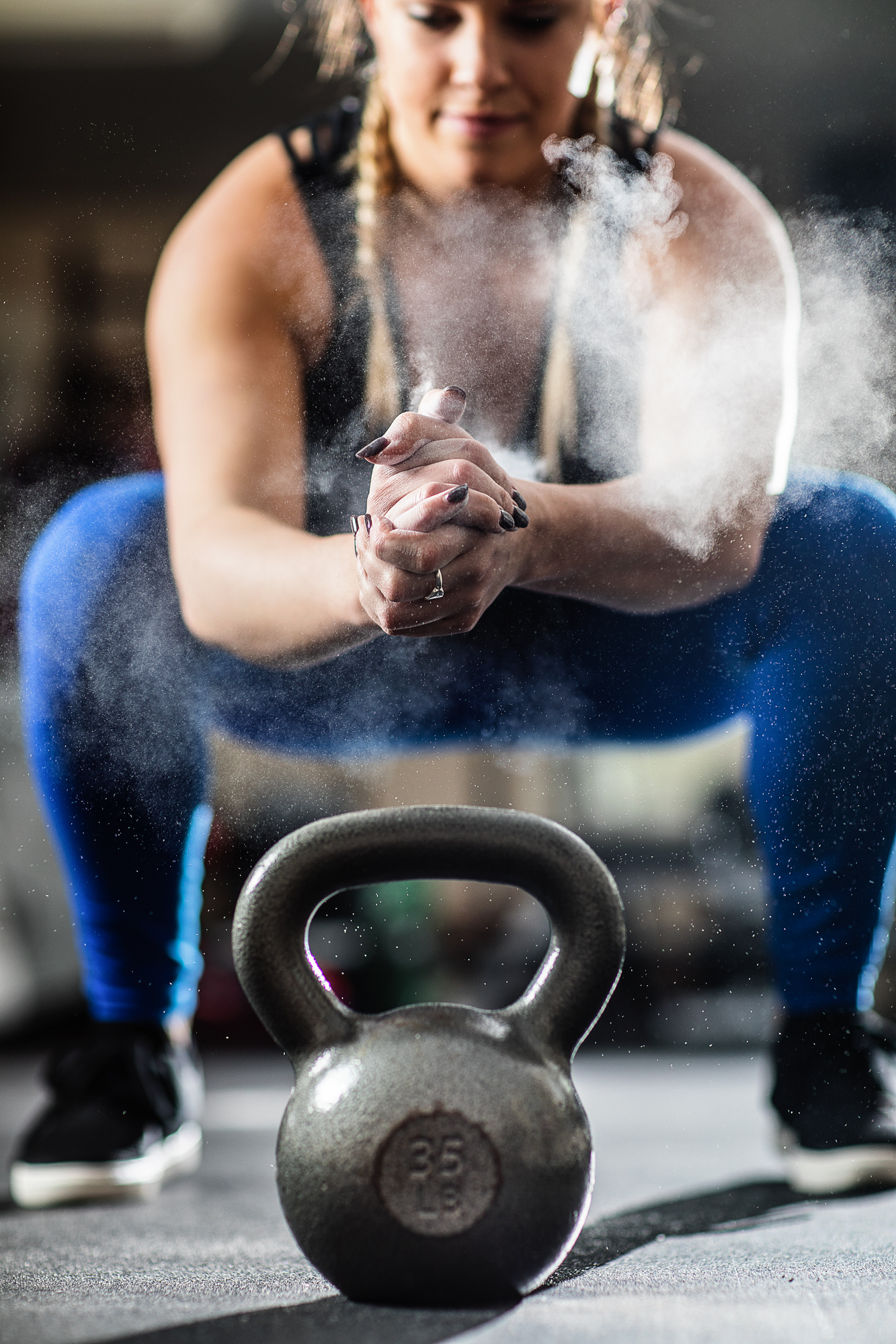 A woman in workout clothes and braids is doing a squat in a gym, with a kettlebell in the foreground and chalk dust around her hands.