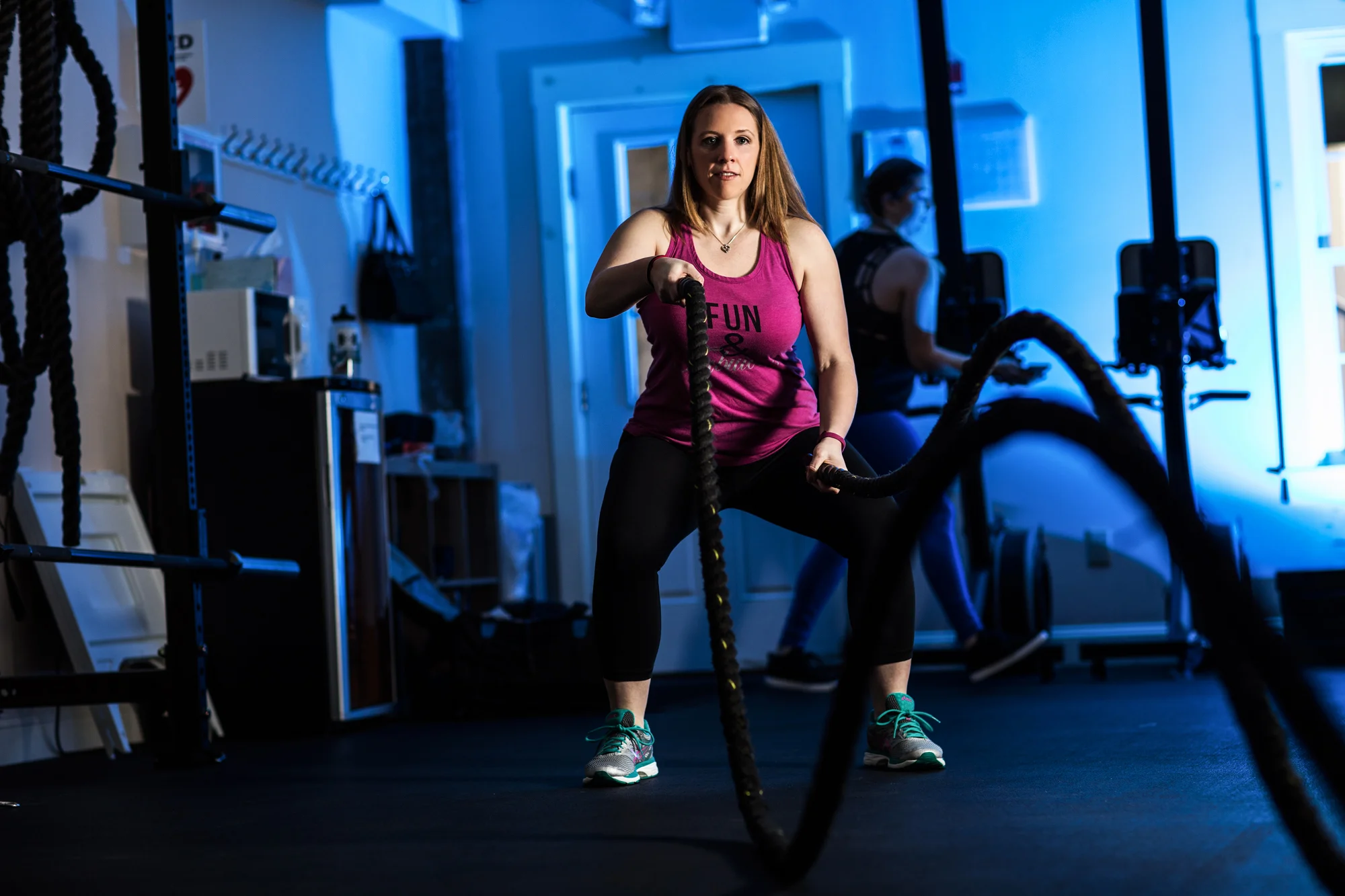 A woman in a pink tank top and black leggings is exercising with battle ropes in a gym. She is in a wide stance and holding the ropes, with gym equipment and another person working out in the background.