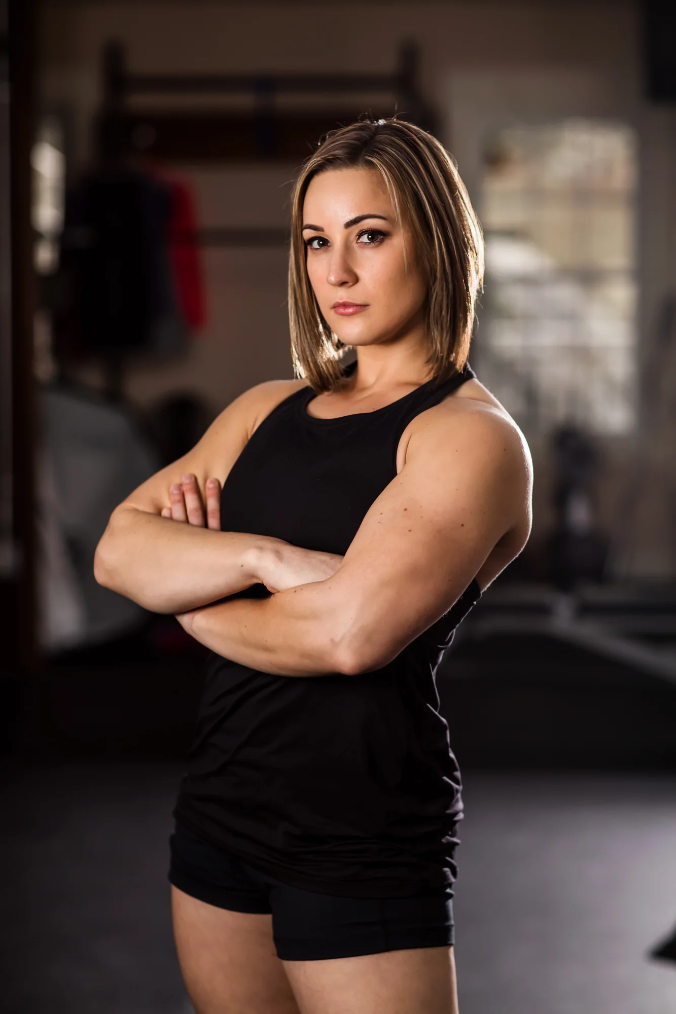 A confident young woman with shoulder-length brown hair standing with arms crossed inside a gym, wearing a black sleeveless workout top and black shorts.