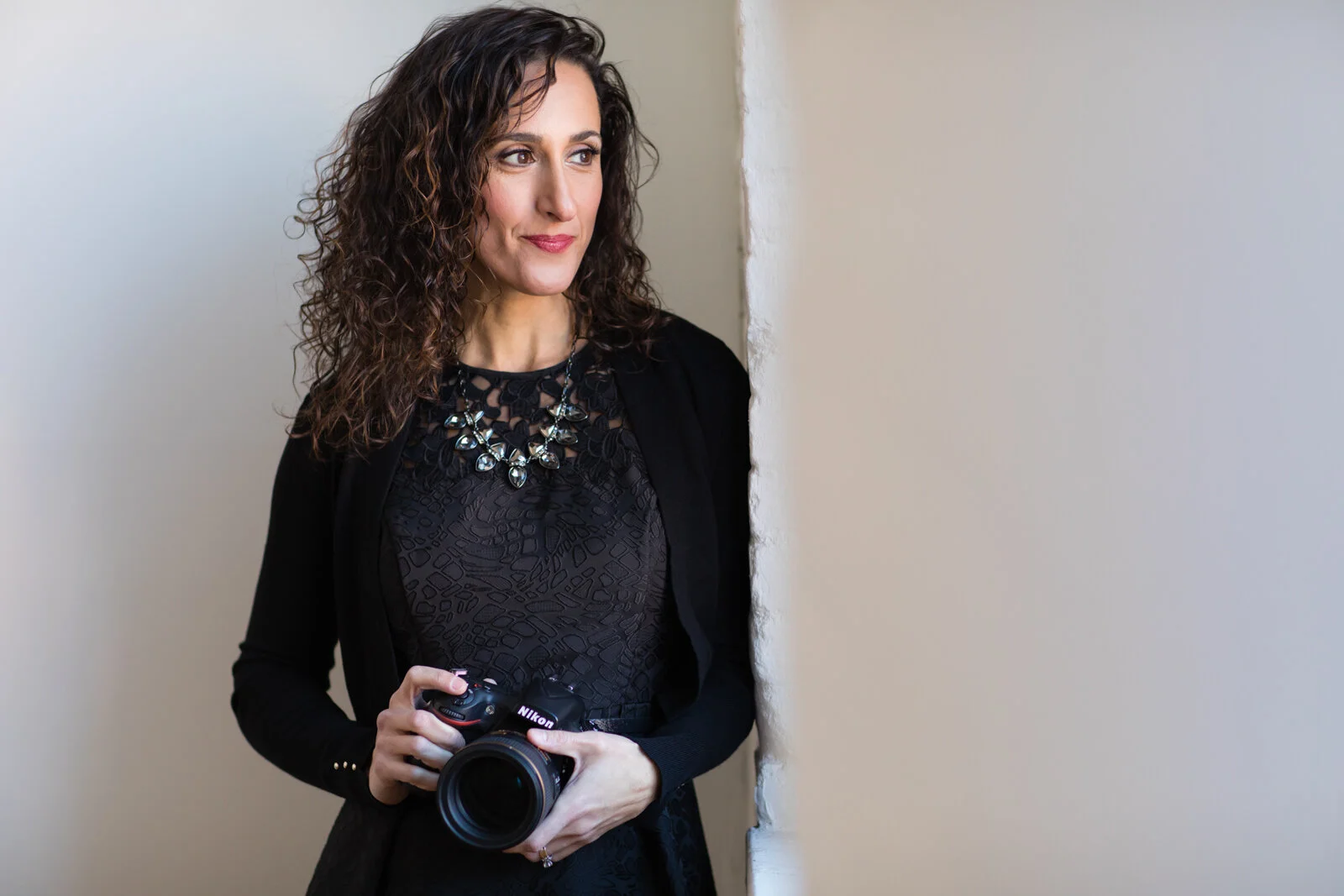 A woman with curly brown hair wearing a black dress and a statement necklace, holding a Nikon camera, standing against a wall.
