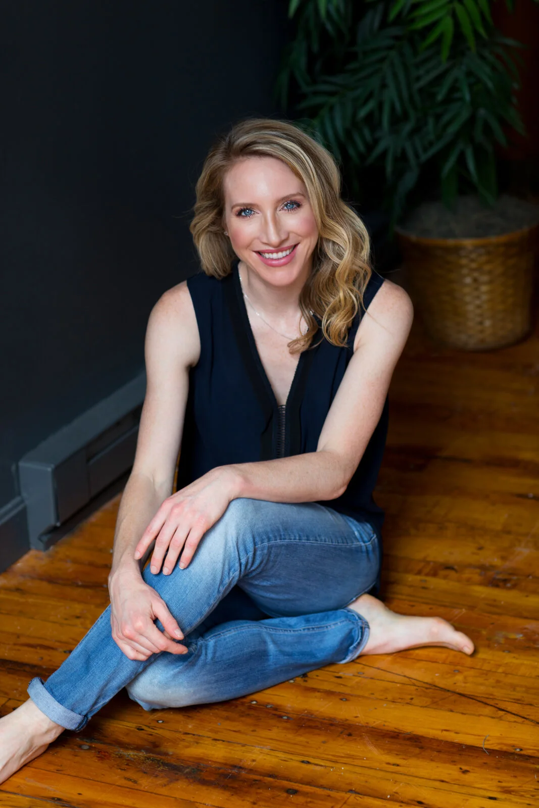 A woman with blonde wavy hair sitting cross-legged on a hardwood floor, smiling, wearing a navy sleeveless top and blue jeans, with a potted plant in the background.