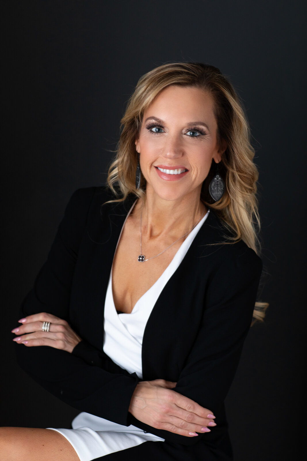 A professional woman with blonde hair, wearing a black blazer over a white top, with jewelry including earrings, a necklace, and rings, smiling against a dark background.