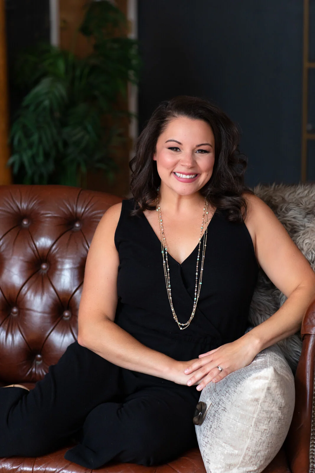 A woman with dark, shoulder-length hair wearing a black sleeveless top and layered necklaces, sitting on a leather couch in a cozy, stylish interior with green plants in the background, smiling at the camera.