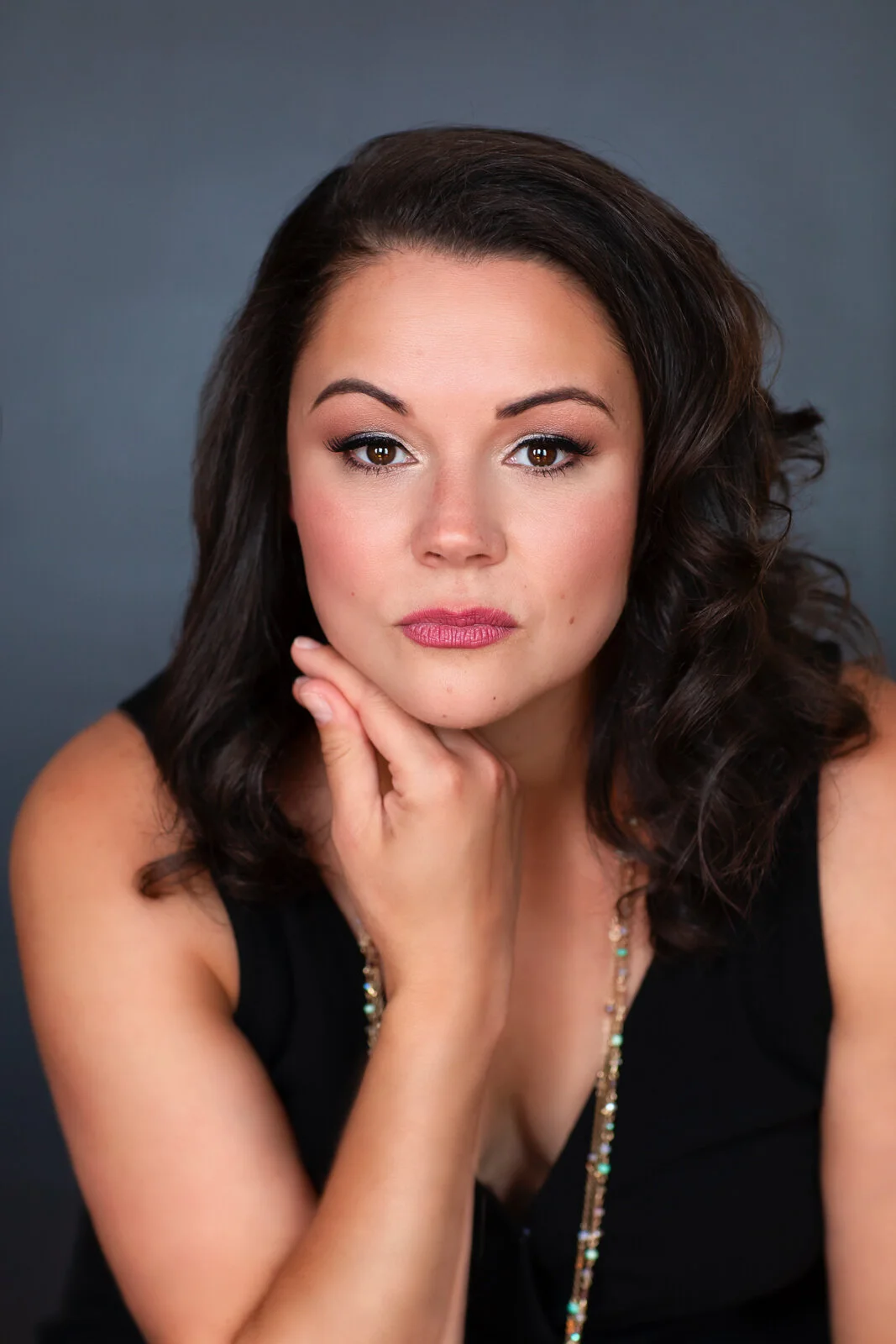 Close-up portrait of a woman with dark brown hair, wearing makeup, a black top, and a beaded necklace, resting her chin on her hand against a dark gray background.