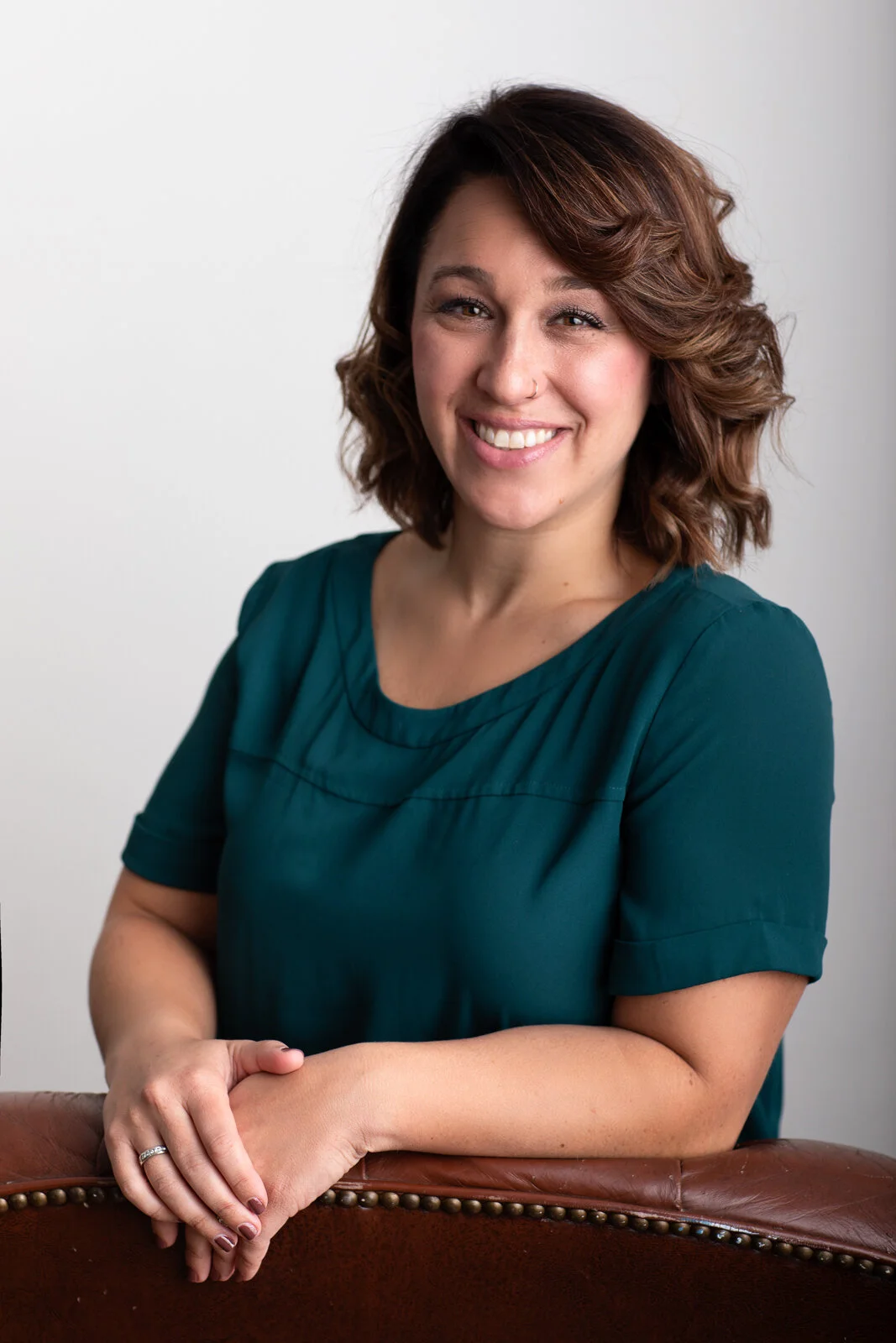 A woman with shoulder-length wavy brown hair, wearing a teal short-sleeved blouse, smiles while leaning on a brown leather surface against a plain white background.