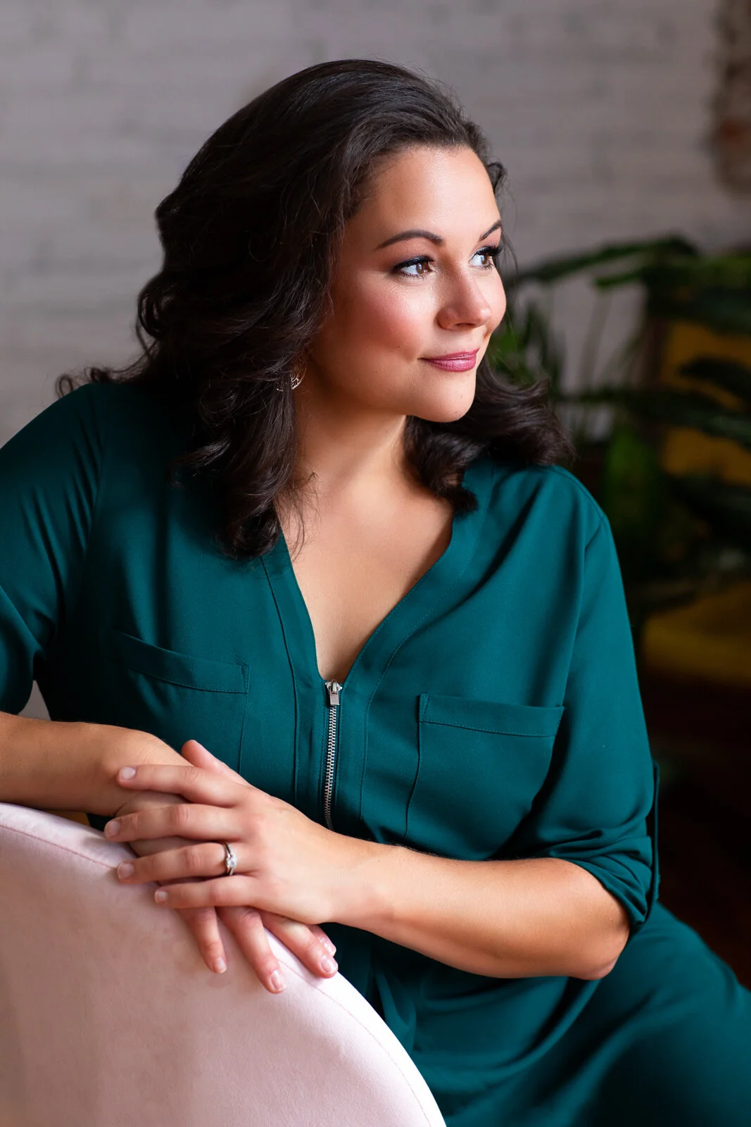 A woman with dark, curly hair sitting and looking to the side. She is wearing a teal dress with a zipper and has a ring on her left hand. The background includes a white brick wall and a plant.