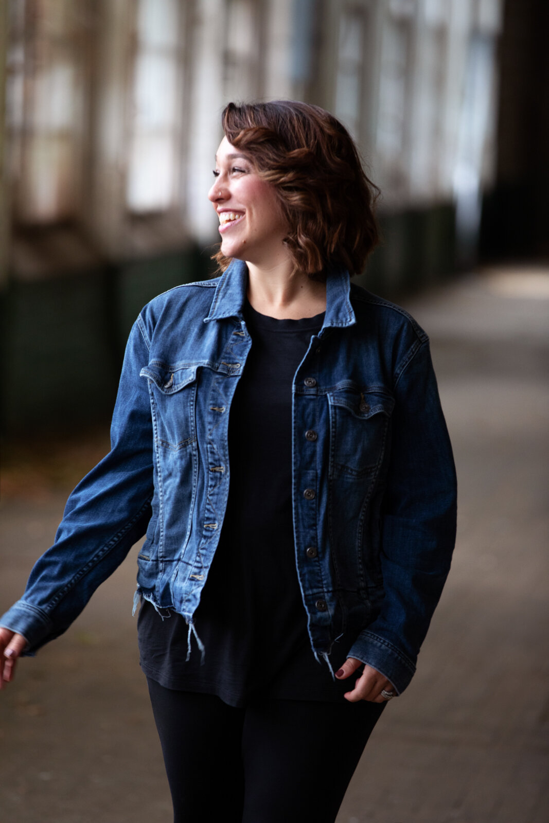 A woman with brown hair wearing a denim jacket and black shirt, smiling and looking to her left, walking in an indoor space with large windows and a blurred background.
