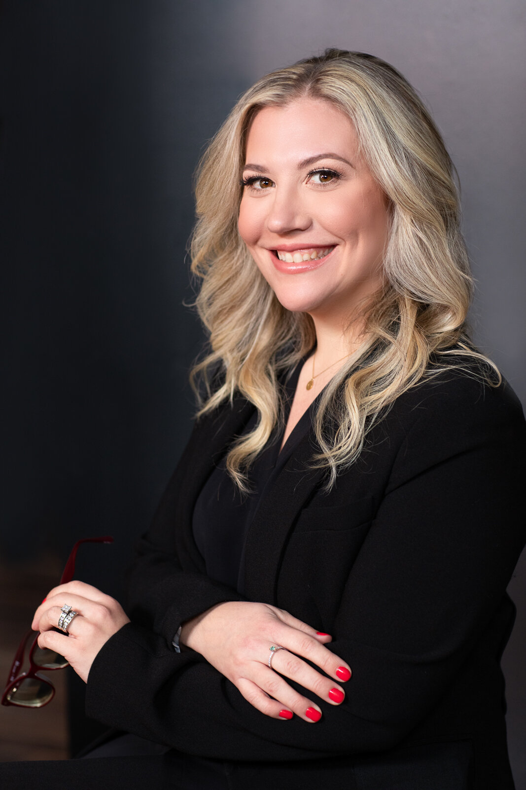 Portrait of a smiling woman with blonde wavy hair, wearing a black blazer, red nail polish, and holding red glasses, against a dark background.