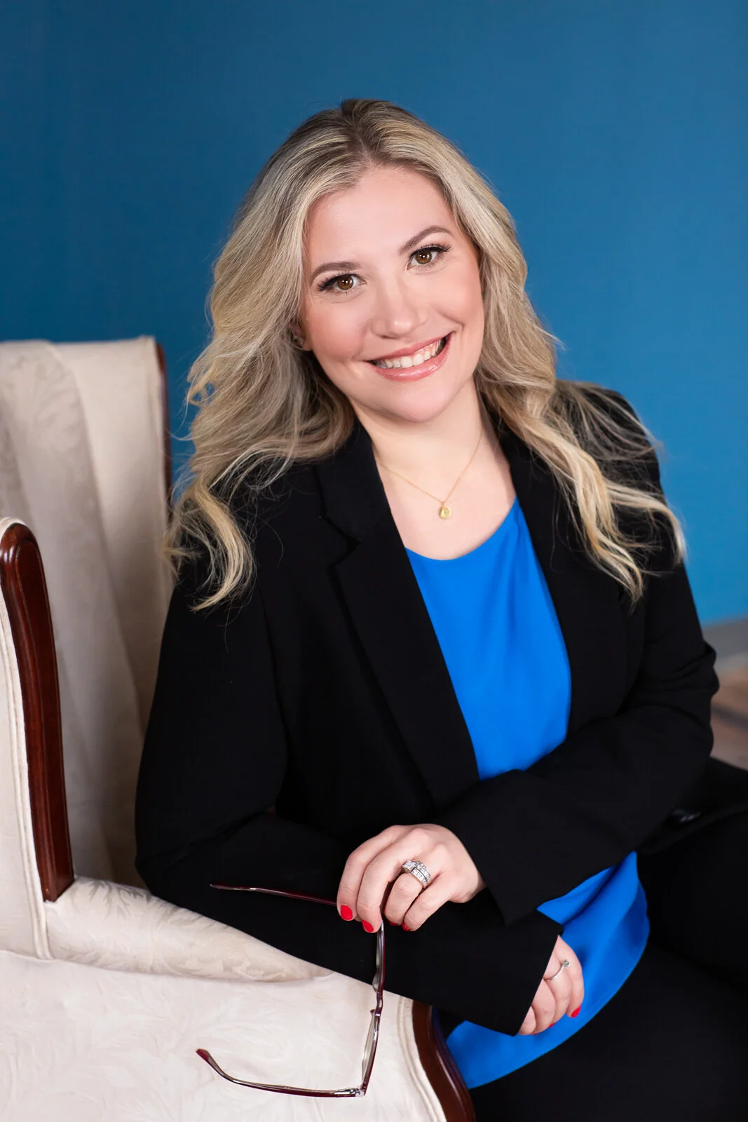 A smiling woman with blonde hair wearing a black blazer and blue top, holding glasses, sitting on a cream-colored chair against a blue background.