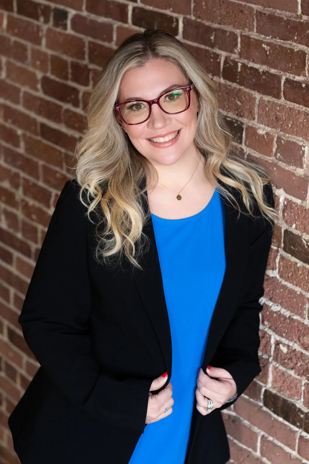 A woman with blonde wavy hair wearing red glasses, a black blazer, and a blue top, smiling and standing against a brick wall.