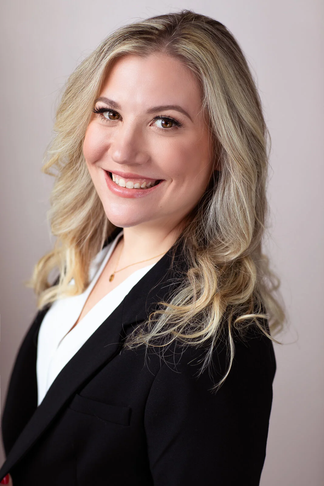 Professional woman with blonde wavy hair, smiling, wearing a black blazer and white shirt, against a neutral background.