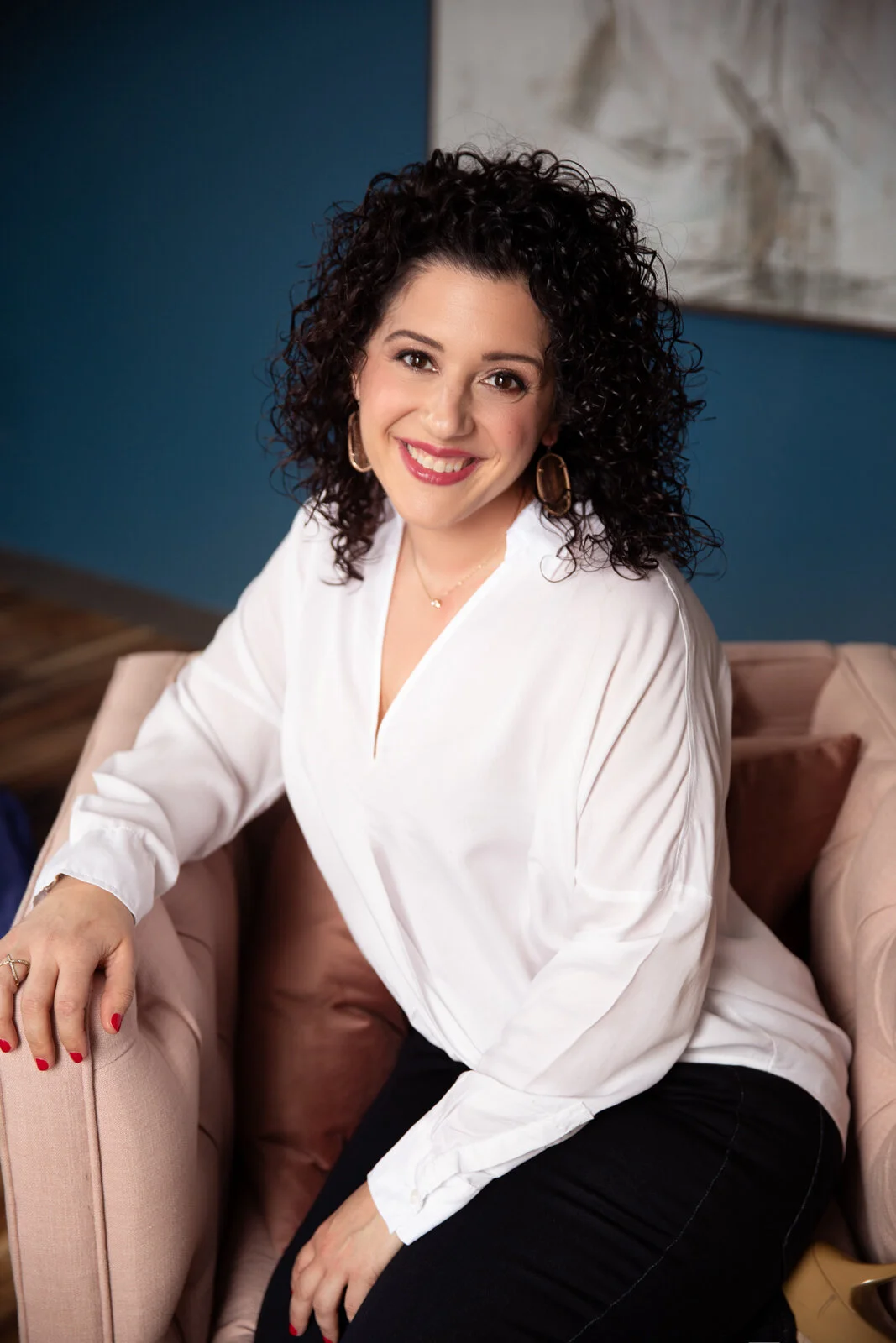 A woman with curly dark hair and a white blouse sitting on a light pink chair and smiling at the camera.