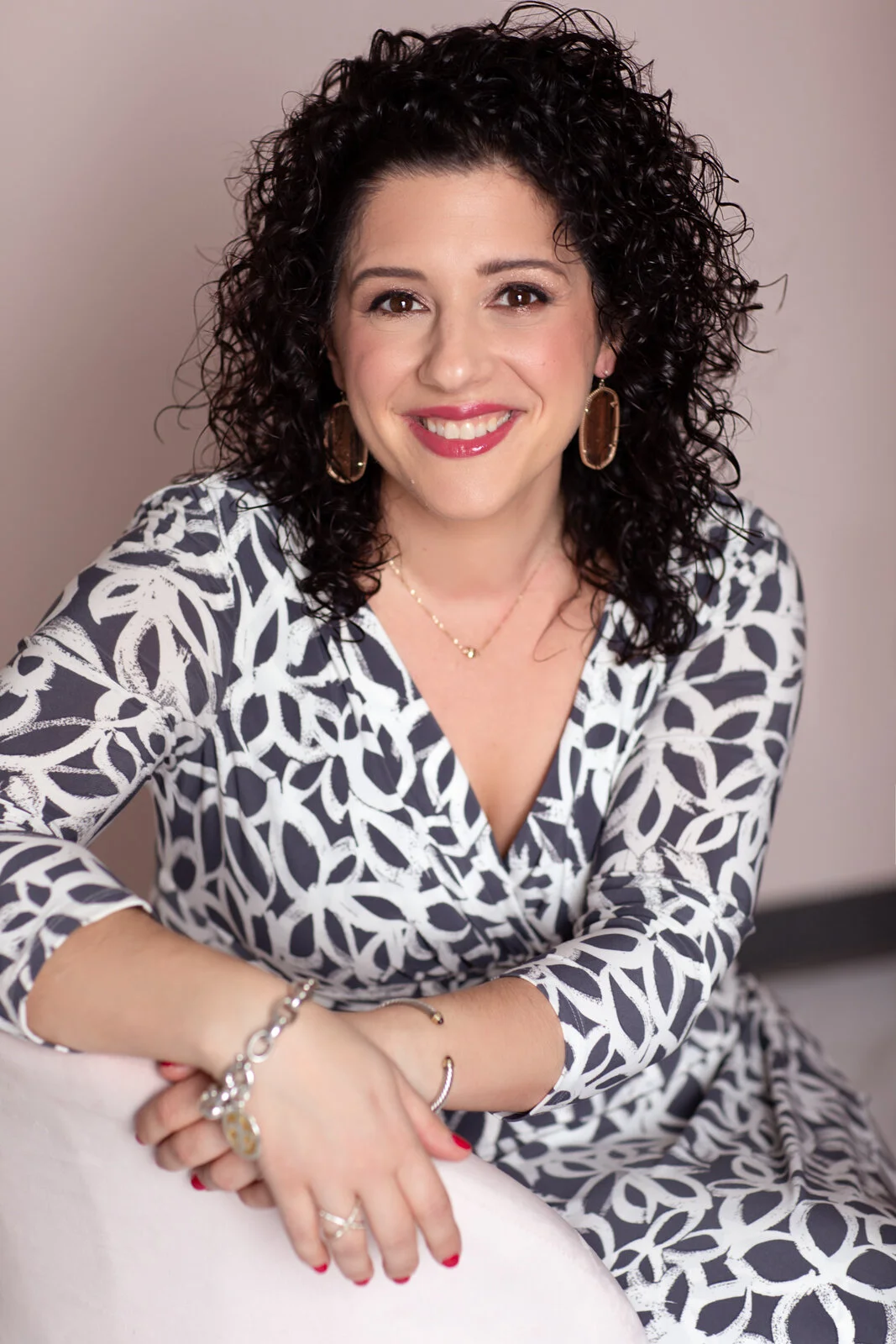A woman with dark, curly hair and light skin, smiling, wearing a black and white patterned dress, gold hoop earrings, and jewelry, sitting against a plain background.