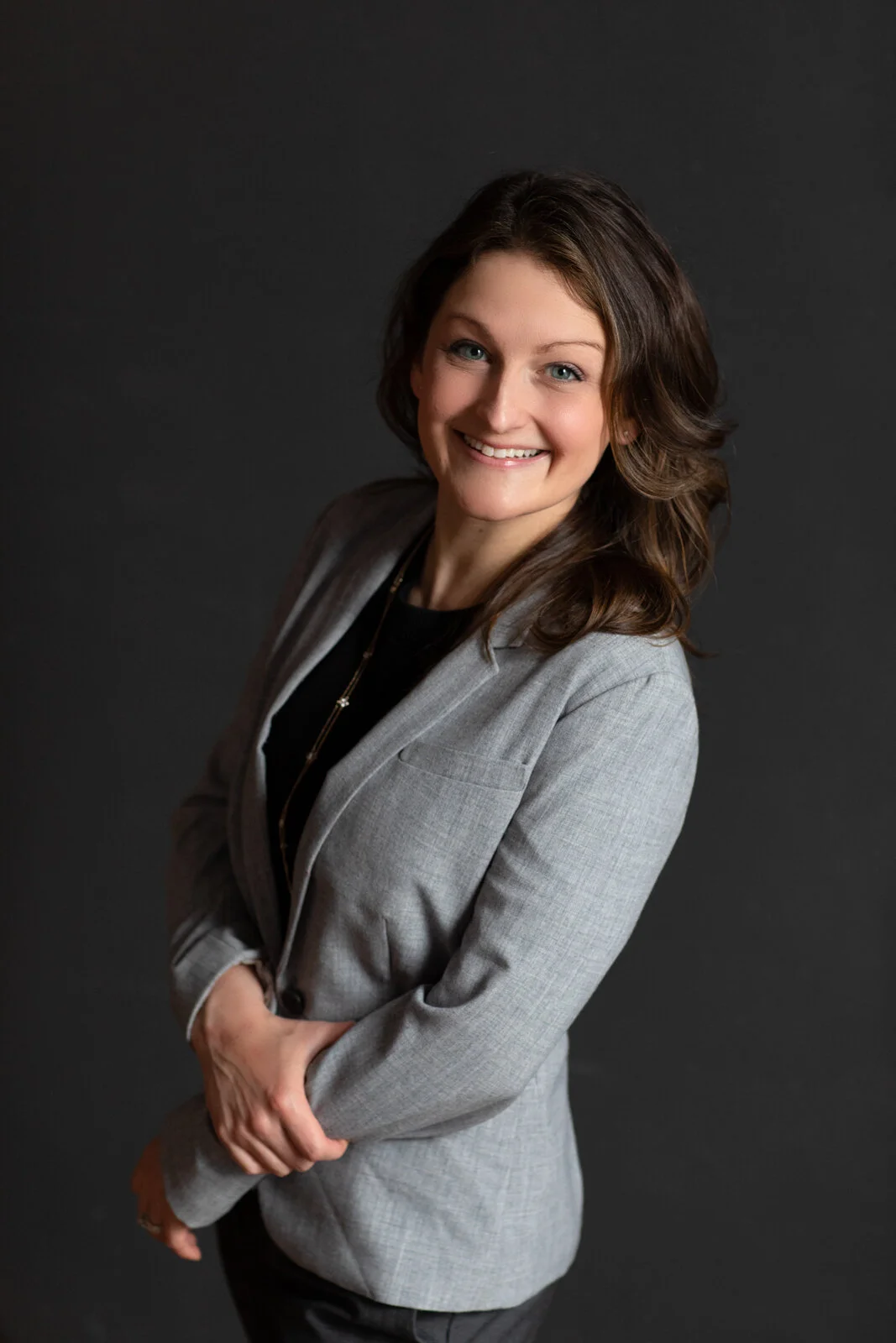 A young woman with long brown hair and blue eyes smiling, wearing a light gray blazer and black top, standing against a dark background.