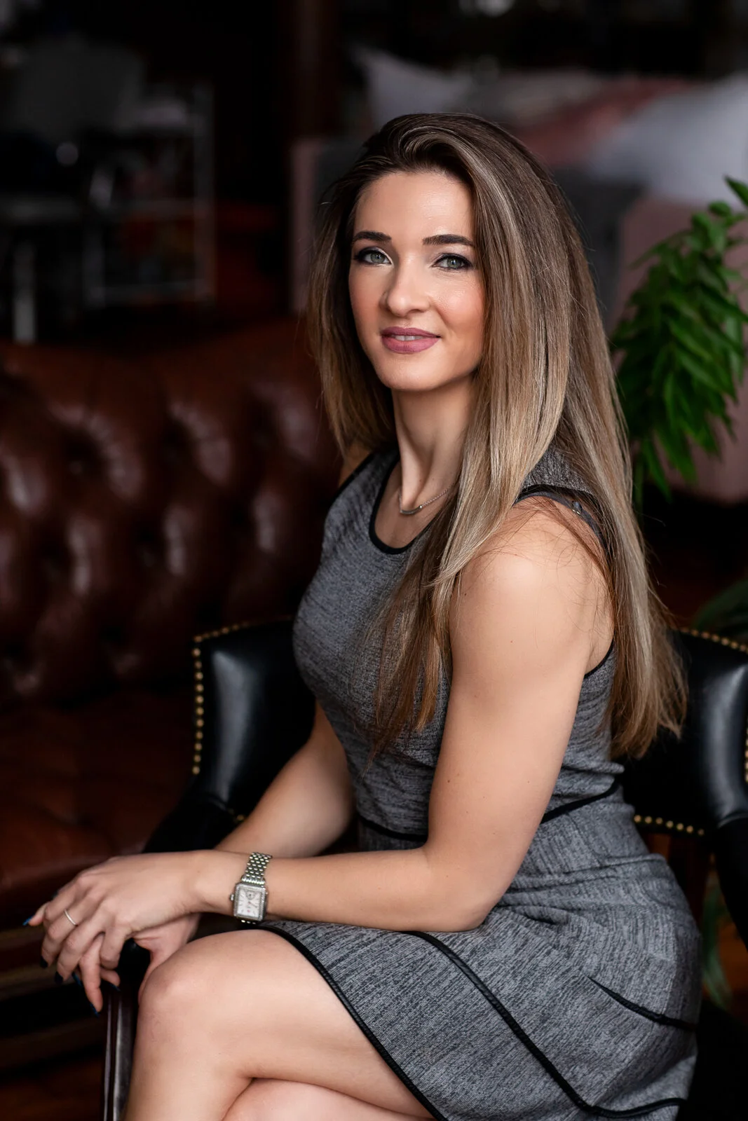 A woman with light brown hair, blue eyes, and fair skin, wearing a sleeveless gray dress with black trim, sitting on a black leather chair inside a room with a leather sofa and green plant in the background.